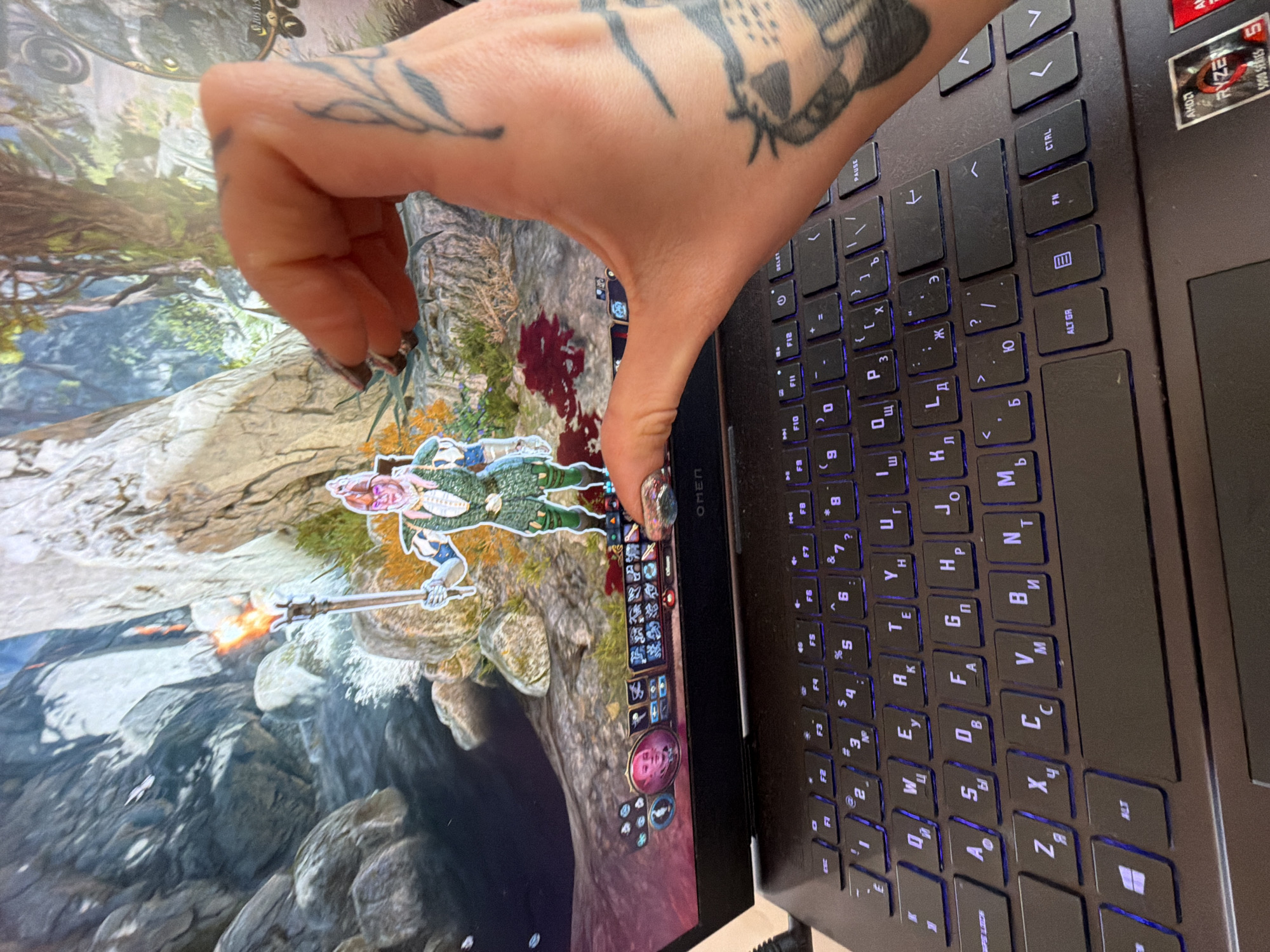 A close-up, slightly angled view shows a person's hand with tattoos over a laptop playing a video game. The hand is positioned over the laptop screen and keyboard. The laptop is an HP Omen with a black keyboard featuring Cyrillic characters. The screen displays an action scene from a video game with characters in a fantasy-style setting. The laptop is an HP Omen with a black keyboard featuring Cyrillic characters. The AMD Ryzen 5000 Series logo is visible on the upper right corner of the laptop's base. The game features a fantasy setting with a character holding a weapon. The hand has nail polish. The location in Budva, Montenegro, cannot be determined from the image.