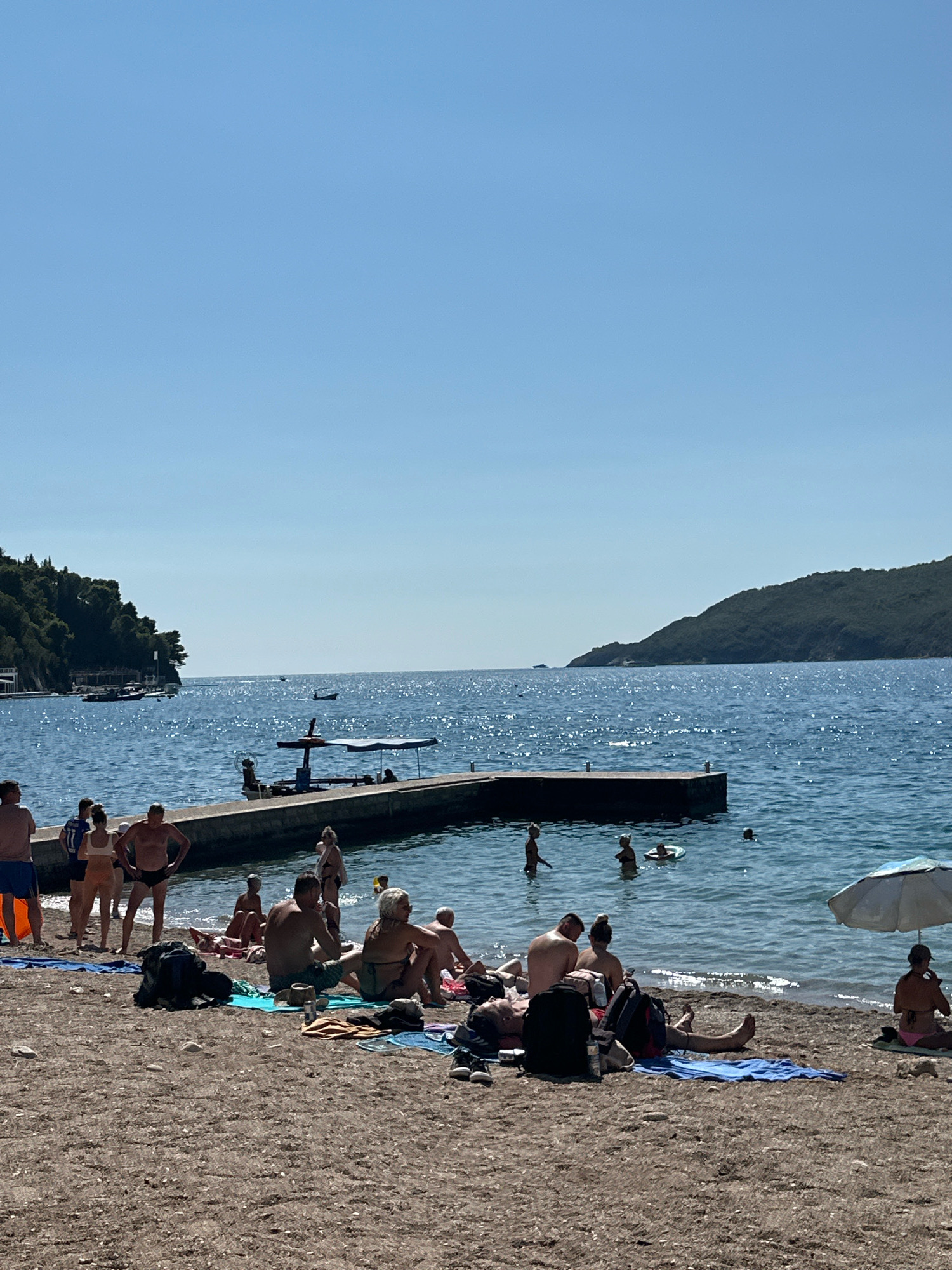 A sunny day at a beach in Budva, Montenegro, shows a shoreline filled with people enjoying the water and sand. The water is calm and blue, with a concrete pier extending into it. Several individuals are swimming or wading in the sea, while others are relaxing on the beach, sunbathing or sitting on towels. Some are engaged in conversation, while others appear to be lost in thought. The beach is made of small pebbles and sand, with a few scattered bags and towels. Lush green hills form a backdrop to the scene, with some buildings visible in the distance. The sky is a clear, bright blue, indicating fair weather.