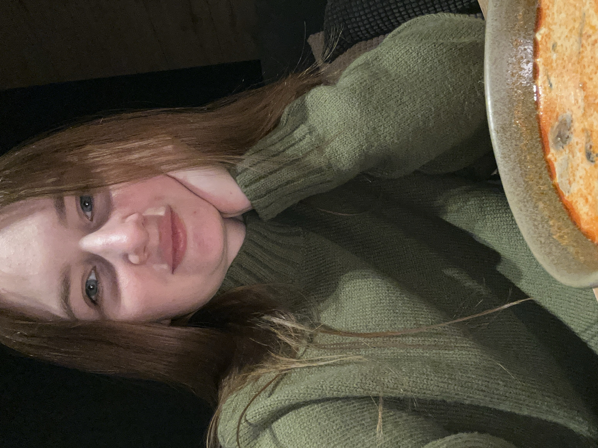 A young woman with long, reddish-brown hair and light eyes is featured in a close-up, dimly lit portrait. She is smiling faintly and looking directly at the viewer. Her left hand is casually placed near her chin and cheek. She is wearing a textured, olive-green knit sweater. To the right of the frame, a ceramic bowl is partially visible, containing an orange-hued dish, possibly a soup or stew, with darker ingredients such as mushrooms or vegetables. The background is dark and out of focus, indicating an indoor setting, possibly a cozy home environment. The low lighting suggests it is evening or night. The image is oriented sideways, with the woman appearing to lie down or lean at an angle. There is no visible text. This scene is set in Chaikovsky, Russia.