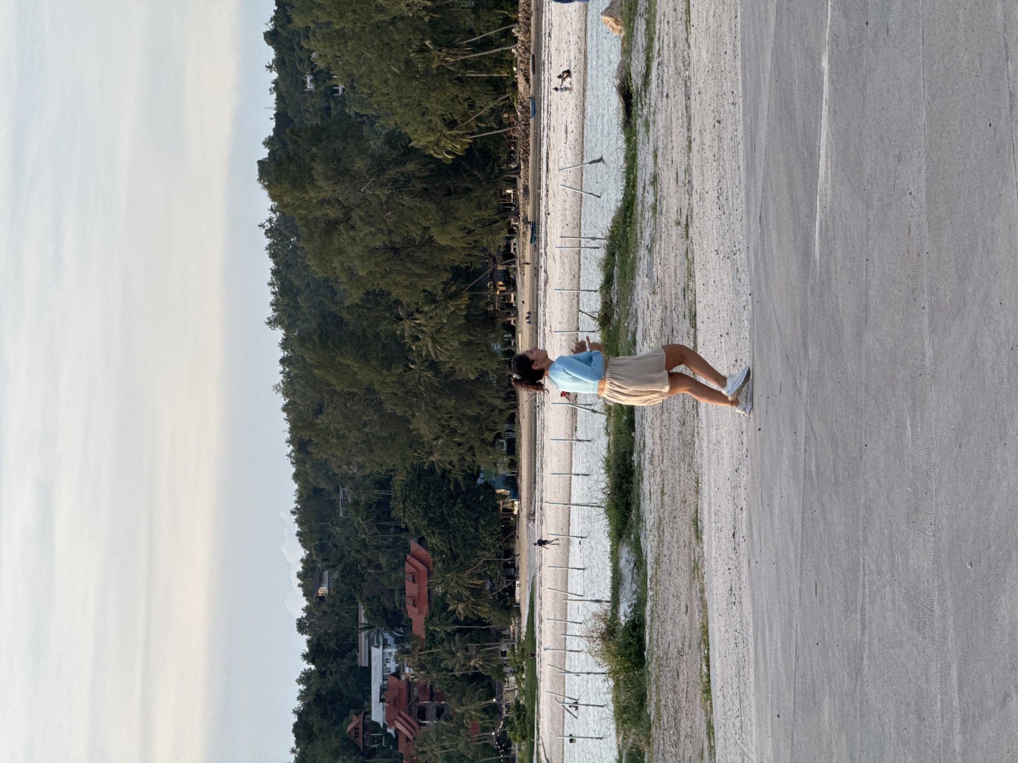 A young woman stands on a paved path, looking out towards a beach. She is wearing a light blue long-sleeved shirt and a tan skirt. The beach is lined with palm trees and lush green foliage. Buildings, possibly hotels or resorts, are visible nestled among the trees. The sand on the beach appears damp, and there are several white poles planted in the sand, spaced evenly apart. The sky is overcast with a pale, diffused light, suggesting it might be early morning or late afternoon. The scene appears peaceful and serene. The woman's pose suggests contemplation or relaxation. The image appears to be taken at a tropical beach location, given the palm trees and the style of the buildings. The location context indicates this is Koh Phangan, Thailand.