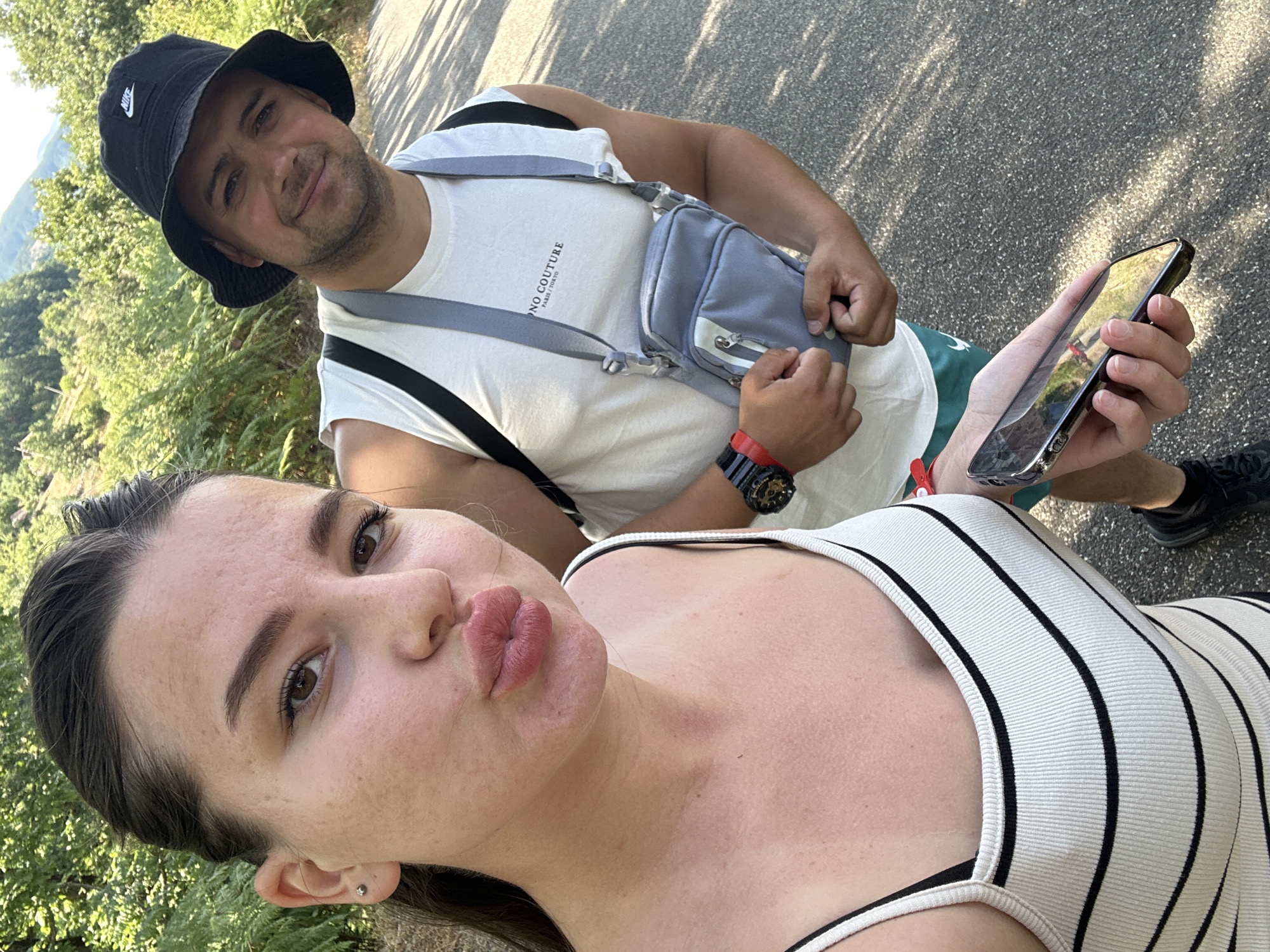 A young woman in a white and black striped tank top is taking a selfie outdoors, puckering her lips towards the camera. She has dark hair pulled back and is wearing a small silver earring. Beside her, a man in a white sleeveless shirt and a black bucket hat smiles at the camera. He is wearing a grey crossbody bag and a red and black watch. The background shows a wooded hillside with green foliage and a paved path. The setting appears to be a natural environment, potentially a park or hiking trail. No specific landmarks within the city of Kremen, Bulgaria, are discernible in the image.