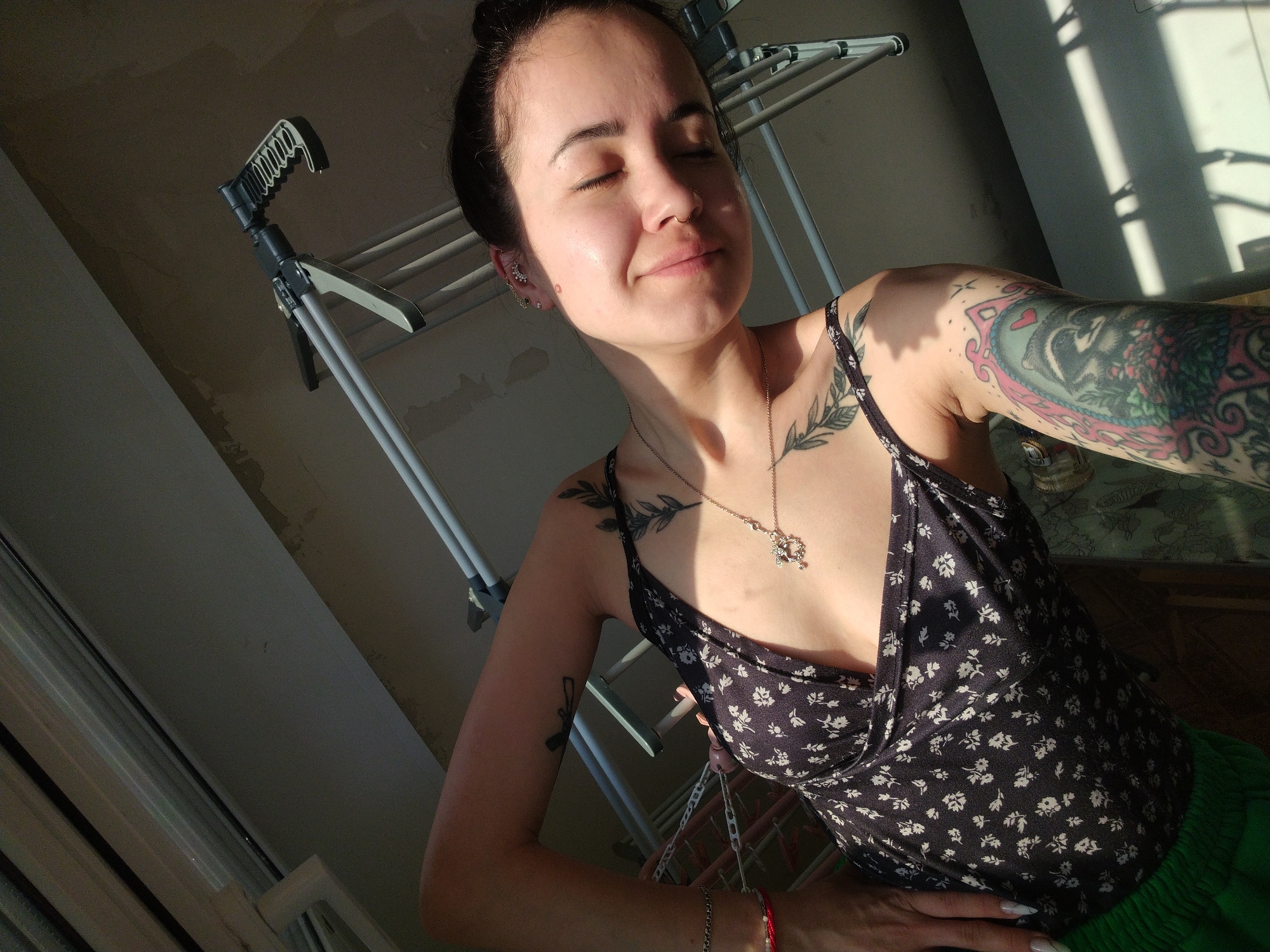 A woman with dark hair pulled back into a bun is posing indoors. She is wearing a black top with a white floral pattern and green bottoms. She has multiple tattoos on her arms, chest, and shoulder. She is wearing a necklace and bracelets. She has her eyes closed and is smiling slightly. A clothes drying rack is behind her. Light from a window is casting shadows on the wall and floor. The location is Noviy, Russia.