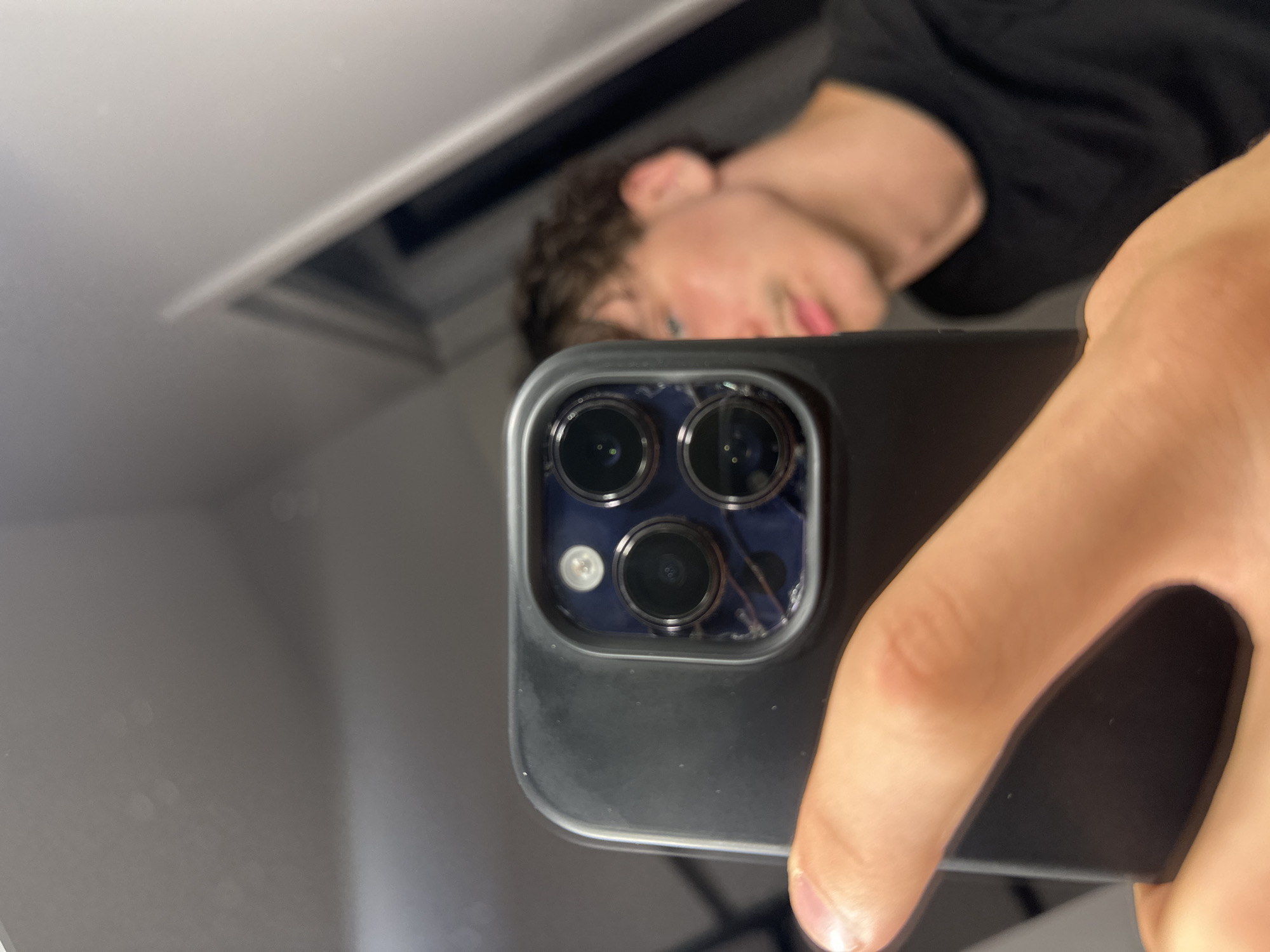 A young person is taking a selfie in a mirror. The image is taken from the perspective of the person holding the phone. The phone's rear camera array, featuring multiple lenses and a flash, is prominently in the foreground. The person's hand is visible holding the phone. In the reflection, the person's face is partially visible, with their gaze directed towards the camera. They are wearing a dark t-shirt. The background is blurred, but appears to be an indoor setting with some light sources. There is no information within the image to suggest the location is in Anduze, France.