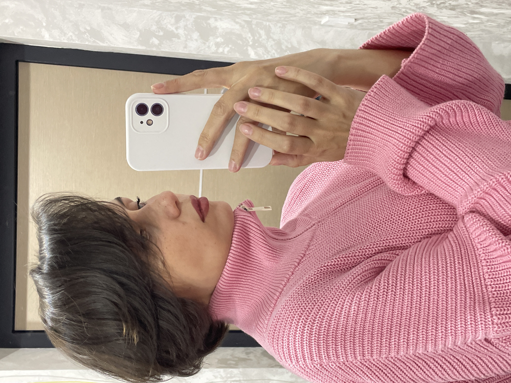 A woman is taking a selfie in front of a mirror. She is wearing a pink, knitted turtleneck sweater. She is holding a white smartphone with a dual-lens camera in her hands. Part of a dark framed mirror is visible behind her. The location cannot be determined from the image.