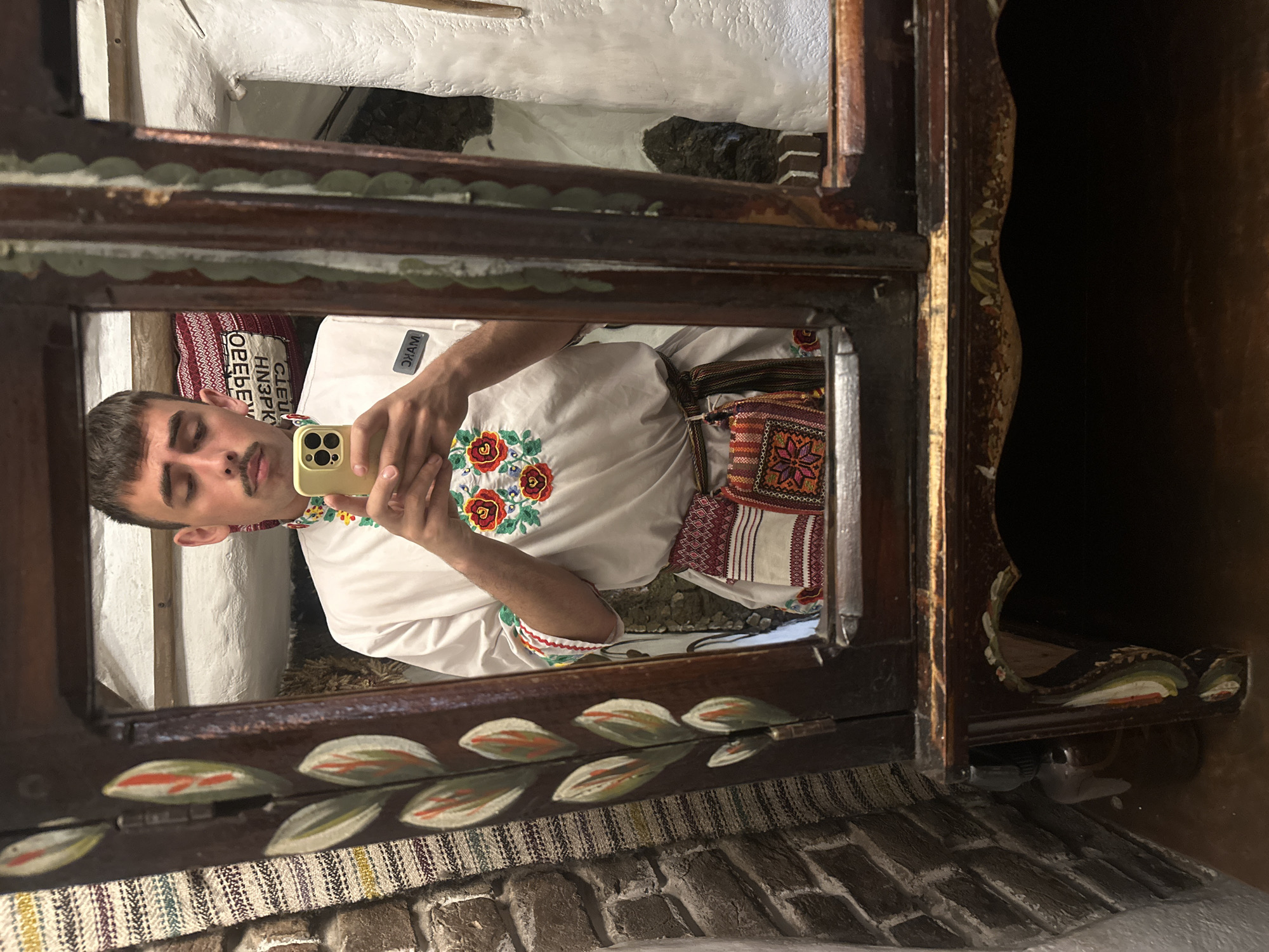 A young man is taking a mirror selfie. He is wearing a traditional white shirt with colorful floral embroidery on the collar and sleeves, a brown belt, and a small embroidered pouch. The man has short, dark hair and a mustache. He is holding a yellow smartphone in his right hand, and it is reflected in the mirror.
The mirror is framed by a dark, ornate wooden cabinet decorated with painted floral motifs. The background in the reflection shows a rustic interior with white walls and some textured fabric. The man appears to be in an indoor setting, possibly a shop or a home in Ternopil, Ukraine, given the context of traditional clothing. The overall scene is well-lit.