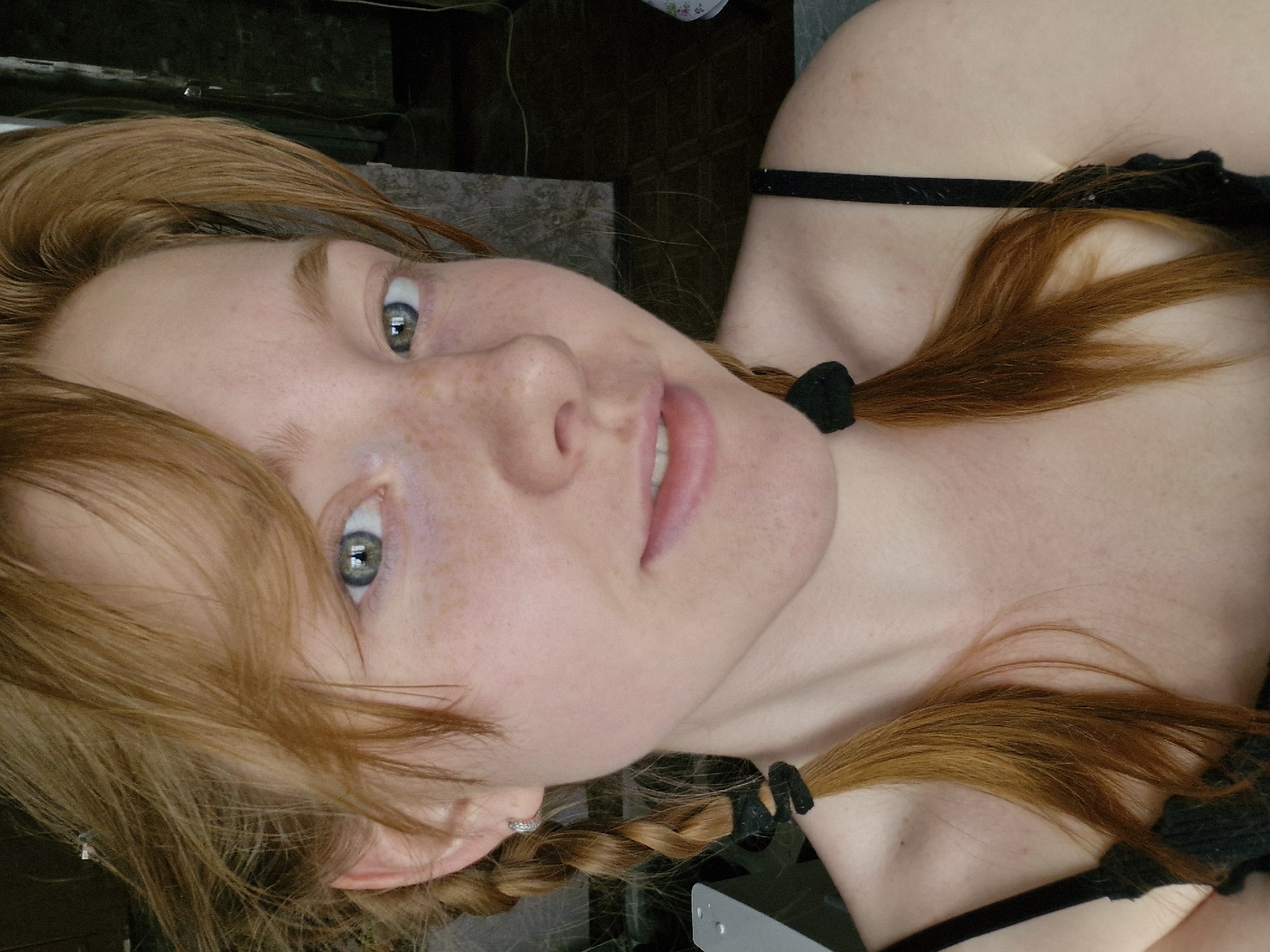 A close-up, slightly tilted portrait captures a fair-skinned woman with light red/ginger hair. Her hair is styled in braids, secured with black bands. She has freckles and gray/blue eyes. She wears a black garment with a strap visible across her shoulder. The background is indistinct, appearing to be a neutral indoor space. There is no evidence to suggest this image was taken in Yekaterinburg, Russia.