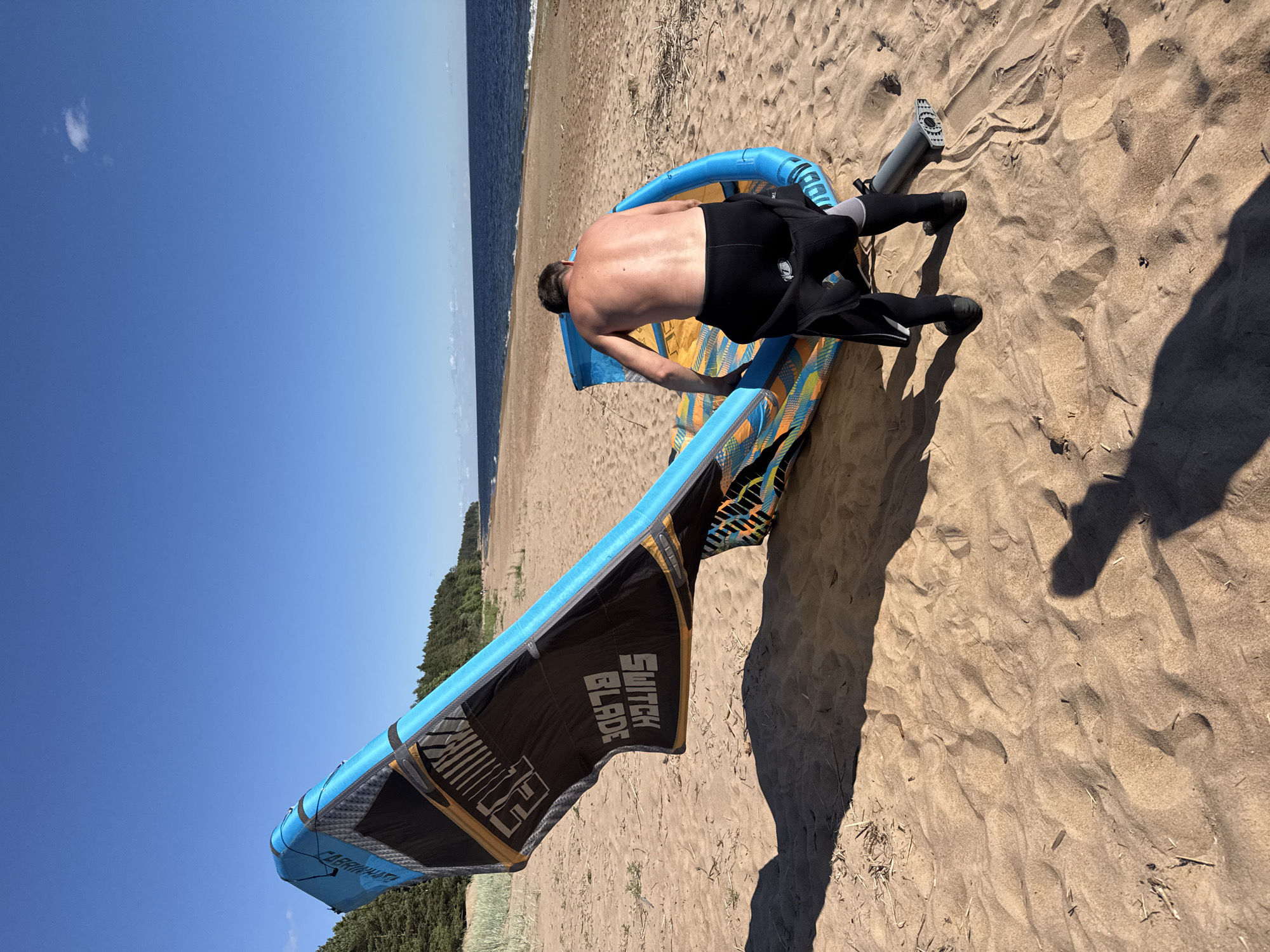 A person wearing a black wetsuit is kneeling on a sandy beach next to a large, blue and orange kite. The kite has the words "SWITCH BLADE" printed on it. The person is facing away from the camera, towards the ocean. The ocean is visible in the background, with a line of trees along the coast. The sky is a clear blue with a few wispy clouds. The sun is high in the sky, indicating it is daytime. The scene appears to be a sunny day at the beach, possibly in Russia, given the "Zelenaya Roshcha" context. The person is likely preparing to kite surf or has just finished.