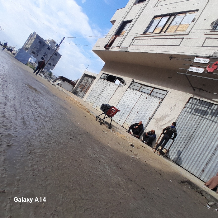 The image depicts a street scene in Khan Yunis, Palestinian Territory, under a partly cloudy sky. The street and adjacent unpaved ground are wet and muddy. On the right, a multi-story building with a light-colored facade exhibits extensive damage, including shattered window panes, boarded-up window openings, and multiple indentations on its metallic roll-down shutters. A red cloth or blanket hangs from an upper-story window. Partially visible signage with Arabic text and phone numbers (e.g., 056877109X, 0597980417) is present on the building's facade.
Three males are situated near this damaged building on the right side of the frame. Two are seated on the dirt ground, leaning against the building's base. A third male stands upright, holding onto a wooden handcart positioned on the ground. A red and black shopping cart rests on the dirt beside the street, near the seated individuals. In the left background, a multi-story concrete structure appears either unfinished or heavily damaged, with exposed rebar and open window frames. A lone individual walks away from the camera on the wet street further down.