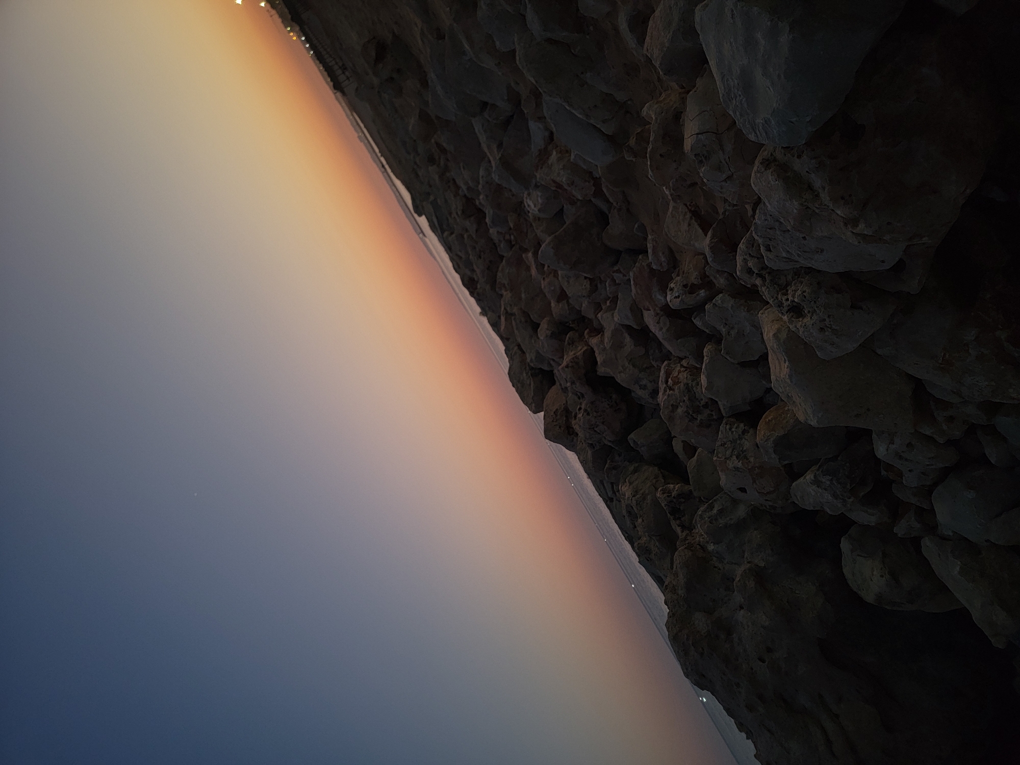 The image depicts a rocky shoreline under a twilight sky. The sky transitions from a deep blue at the top to a gradient of pale orange and pink near the horizon, suggesting either sunrise or sunset. The sea is visible as a dark, calm expanse reflecting the faint light of distant lights along the coast. The foreground is dominated by a jumble of large, rough rocks, casting shadows and creating a textured surface. In the far distance, along the shoreline, faint lights indicate man-made structures, possibly streetlights or buildings. No people or animals are visible in the image. The scene is devoid of any specific identifying landmarks that would pinpoint a location within Quarteira, Portugal, beyond being a coastal area.