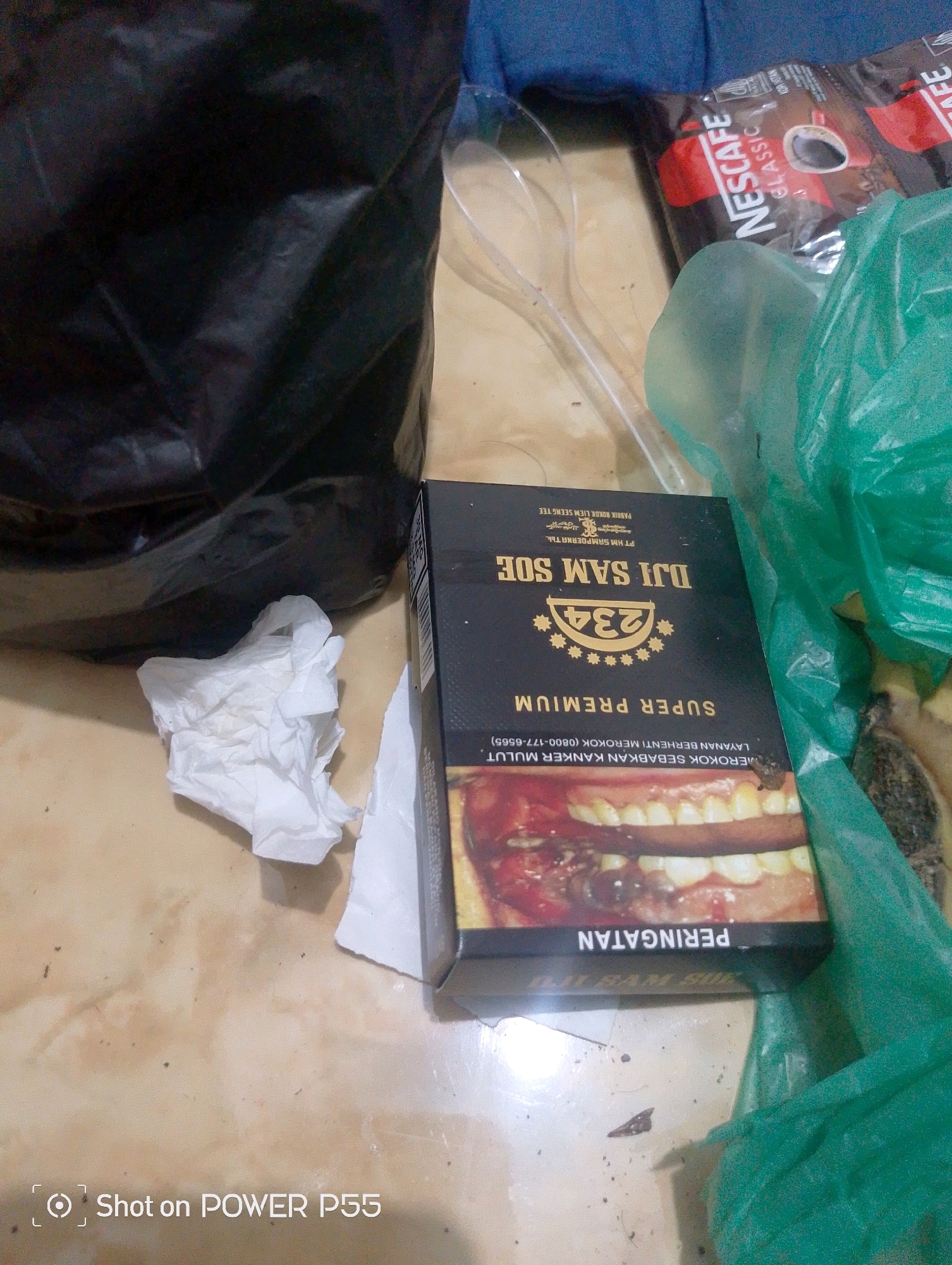 The image shows a close-up of a table surface. In the foreground, a black and gold cigarette pack with the brand "DJI SAM SOE SUPER PREMIUM" is visible. The pack displays a graphic image of a mouth with decayed teeth and gums, along with warning text in Indonesian. To the left of the cigarette pack, a crumpled white tissue is present. Behind the cigarette pack and tissue, a black plastic bag is partially visible. To the right of the cigarette pack, a bright green plastic bag is also partially visible, with what appears to be food items inside. In the background, a packet of "NESCAFE CLASSIC" coffee and two clear plastic spoons are on a blue surface. The floor appears to be tiled, with some dirt and debris scattered on it. A "Shot on POWER P55" watermark is in the bottom left corner. The scene is likely indoors, given the presence of the table and various packaged items. The specific location in Lebaksiu, Indonesia, is not discernible from the image itself.