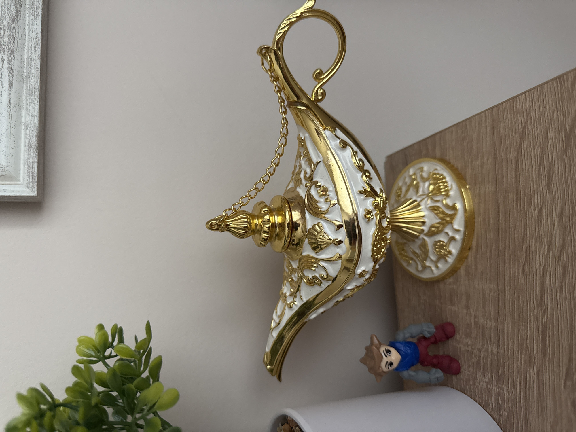 A photograph shows a decorative Aladdin's lamp, white and gold, hanging from a wall. Beside the lamp, there is a small figurine of a boy with brown hair, a blue shirt, and red pants. The background includes a white wall, a faux green plant in a white pot, and a wooden surface. A partially visible white framed picture is on the left. The location in the city of Lovech, Bulgaria is unknown.