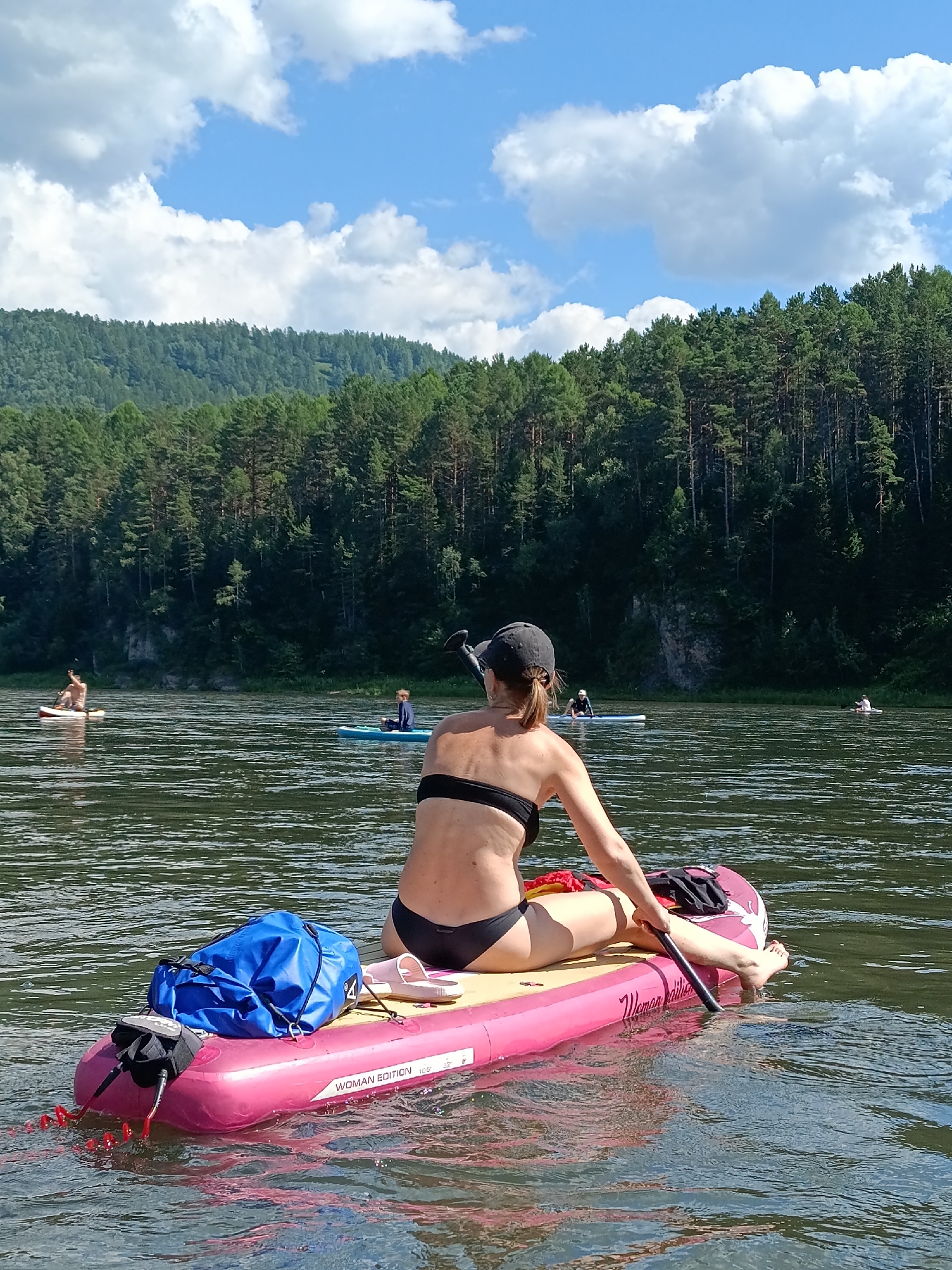 In the city of Ust-Mana, Russia, on a wide, calm river with a slight chop, a woman in a black bikini and black baseball cap sits on a pink inflatable paddleboard. She is facing away from the camera, holding a black paddle with her right hand, her feet submerged in the water. On the paddleboard are a blue waterproof bag and a pair of pink flip-flops. The paddleboard has "WOMAN EDITION 10'8"" printed on its side. In the background, across the river, is a dense forest of green pine trees that covers a sloping hillside, with a bright blue sky and white clouds above. Other individuals on paddleboards are visible further down the river, also surrounded by the forest and water.