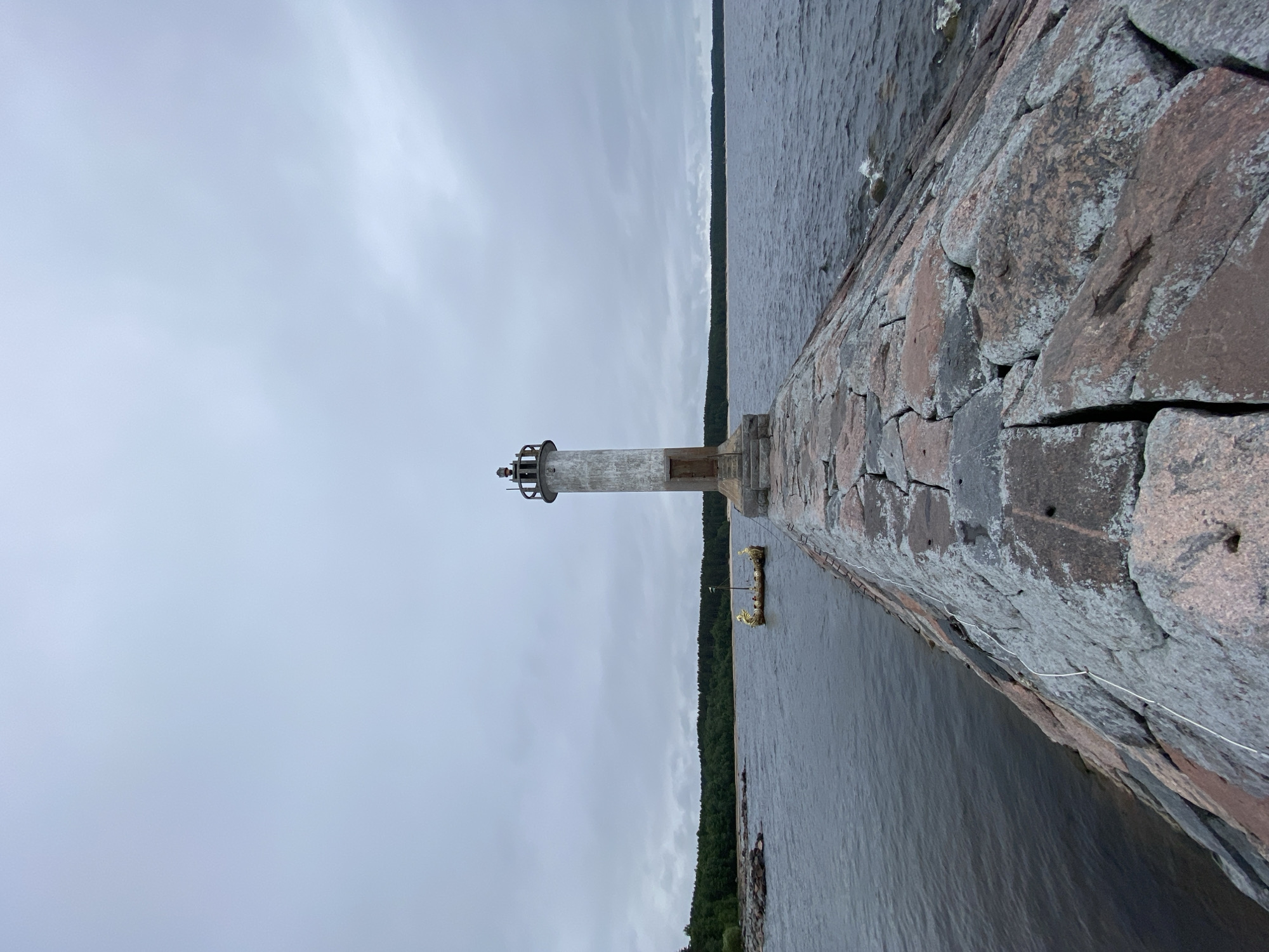 A lighthouse stands on a stone pier extending into dark, choppy water under an overcast sky. The lighthouse is cylindrical and made of concrete with a metallic top. A wooden door is visible at the base of the lighthouse. The pier is constructed from large, rough-hewn stones, some of which are covered in lichen. To the left of the pier, a small, ornate boat with figures and decorative elements is moored. The background features a dense forest lining a sandy shoreline, with more rocky outcrops visible on the left. The overall scene conveys a sense of remoteness and an industrial maritime setting. The location is the lighthouse on the pier in Motornoe, Russia.
