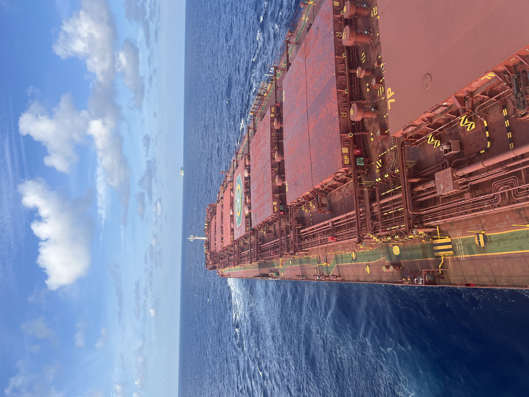 An aerial, high-angle shot captures the deck of a large, red cargo ship sailing through the ocean under a bright blue sky filled with white, puffy clouds. The ship's expansive deck is covered in red panels, with visible yellow markings and numerous pipes and machinery. The turbulent, blue ocean surrounds the vessel, with white foam trailing from its wake. In the distance, two smaller cargo ships are visible on the horizon. The scene is located at an unknown place in an unknown city.