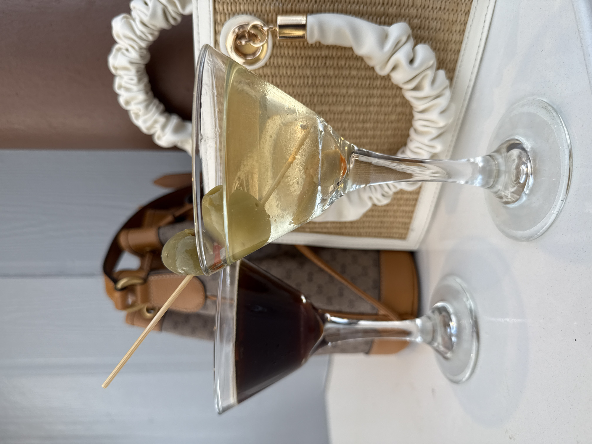 Two martini glasses, one filled with a light-colored liquid containing olives on a toothpick, and the other with a dark-colored liquid, sit side by side. Behind the glasses are two handbags: one white with a distinctive handle design, and the other tan with a patterned logo and brown leather straps. The setting appears to be a flat surface, possibly a table. The image was taken in Orlando, United States of America.