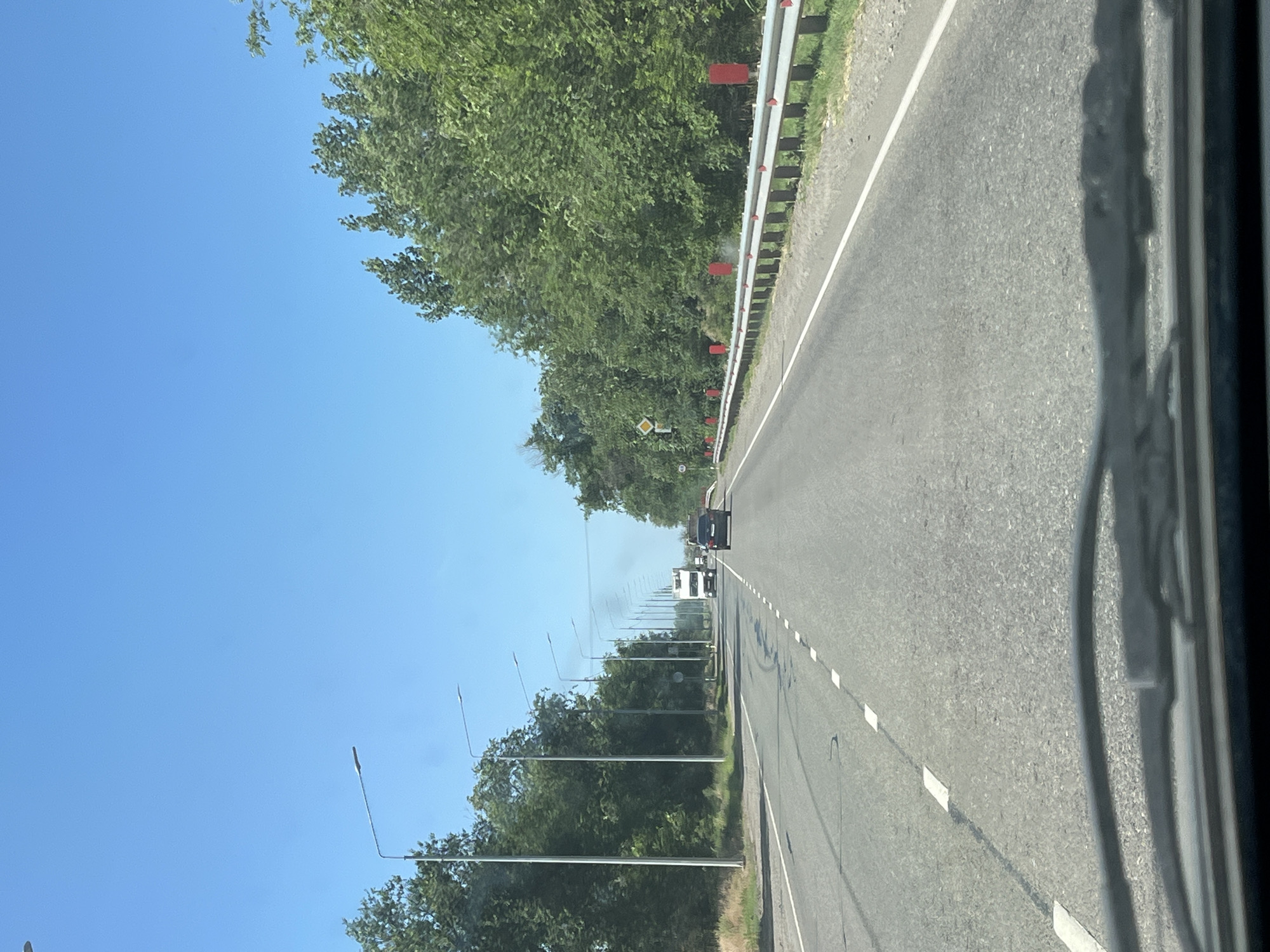 The image depicts a highway scene in Algaza, Russia, under a clear blue sky. The perspective is from inside a vehicle, looking forward through the windshield. A two-lane asphalt road stretches into the distance, flanked by lush green trees on both sides. Streetlights are positioned along the left side of the road, and a metal guardrail with red markers lines the right side. Several vehicles are visible on the road: a white truck is in the distance, followed by a dark-colored car. Smoke is emanating from the truck, suggesting it might be a diesel vehicle. The road is marked with white dashed lines and some darker patches, possibly indicating wear or repairs. The environment appears to be rural or suburban, with no visible buildings.