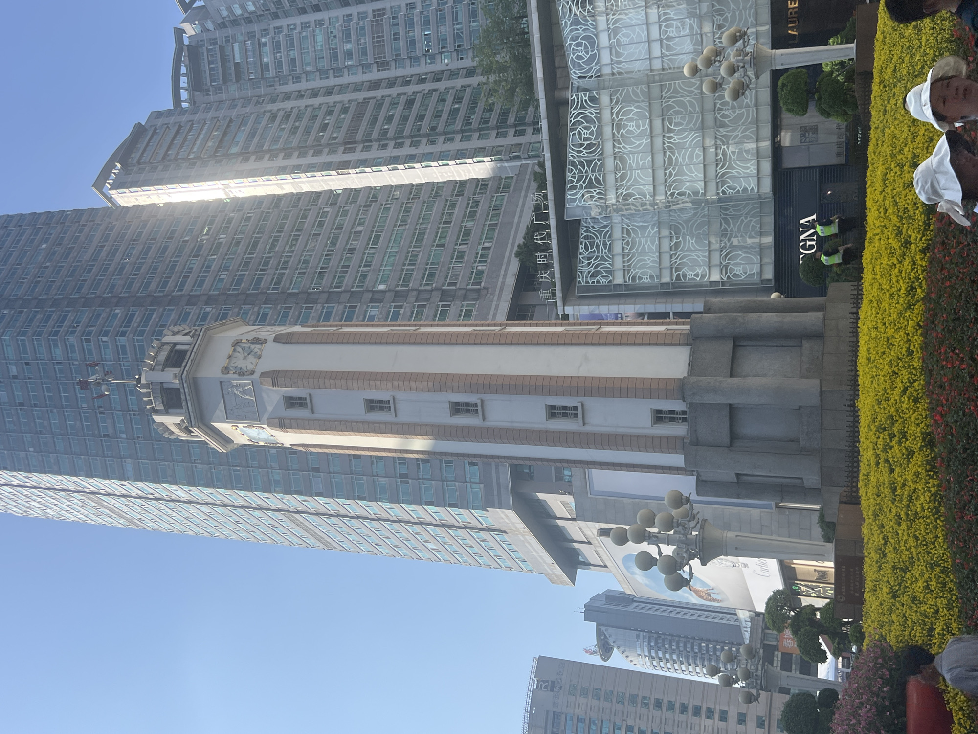 The image captures a bustling urban scene in Chongqing, China, under a bright, clear blue sky. The perspective is upward, emphasizing the height of the surrounding buildings.
In the foreground, a distinctive, ornate tower with a light-colored facade stands out. This structure, which appears historical, features a clock face near its summit and detailed carvings or sculptures. Its base is constructed of darker stone, and narrow rectangular windows punctuate its sides.
Surrounding and towering above this historical element are several modern skyscrapers, predominantly constructed of glass and steel, reflecting the sunlight. One notable modern building exhibits an elaborate, almost floral-patterned glass facade.
At street level, the scene is adorned with meticulously maintained landscaping, including vibrant yellow-green hedges and patches of red flowering plants. Globe-style streetlights are also visible.
Several people are present. Some individuals are seen wearing white hats, possibly tourists or part of a group. Two individuals in high-visibility vests, likely security personnel, are stationed near a building entrance.
Visible text on the buildings and signs provides context. The name "重庆时代广场" (Chongqing Times Square) is prominently displayed on a large building, identifying the location. Other visible business names include "GNA" on a storefront, a partial "LAUREN" (likely part of Ralph Lauren), and a large advertisement featuring the "Cartier" logo.
The scene depicts a dynamic urban environment where traditional architecture harmoniously coexists with modern skyscrapers, suggesting a vibrant commercial and cultural hub during daylight hours.