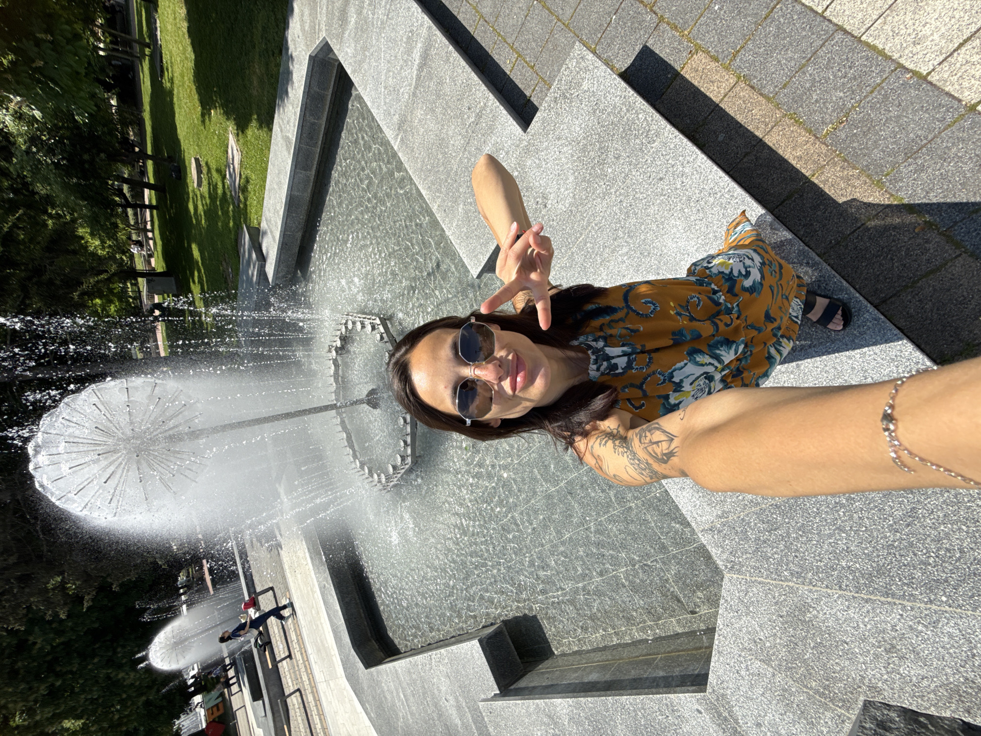A woman is taking a selfie at the Pleven Water Cascade, a fountain in Pleven, Bulgaria. She is wearing sunglasses and a floral dress. The fountain is visible in the background.