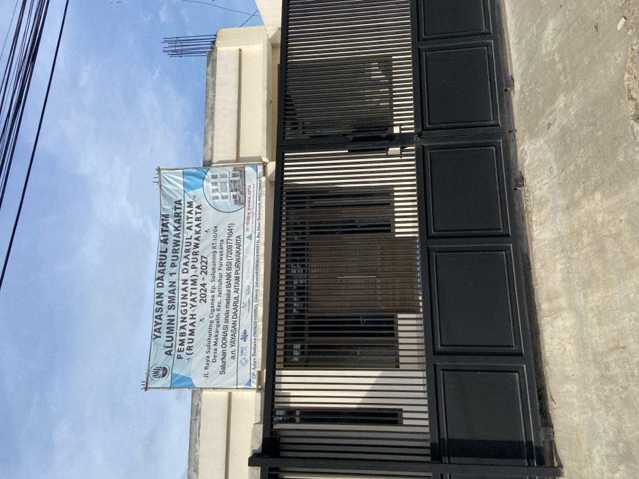 The image shows a black metal gate in front of a building with a banner hanging above it. The banner says "Yayasan Daarul Aitam Alumni SMAN 1 Purwakarta Pembangunan Daarul Aitam (Rumah Yatim) Purwakarta 2024 - 2027" and includes contact information. The gate is made of vertical metal bars and has a decorative design. The gate is closed and there are no people present. The building behind the gate is a two-story structure with a flat roof. The building is painted white and has several windows. The scene is set against a clear blue sky. The image is taken in Jatiluhur, Indonesia. The coordinates -6.571, 107.44, correspond to a location in the city of Purwakarta, Indonesia.