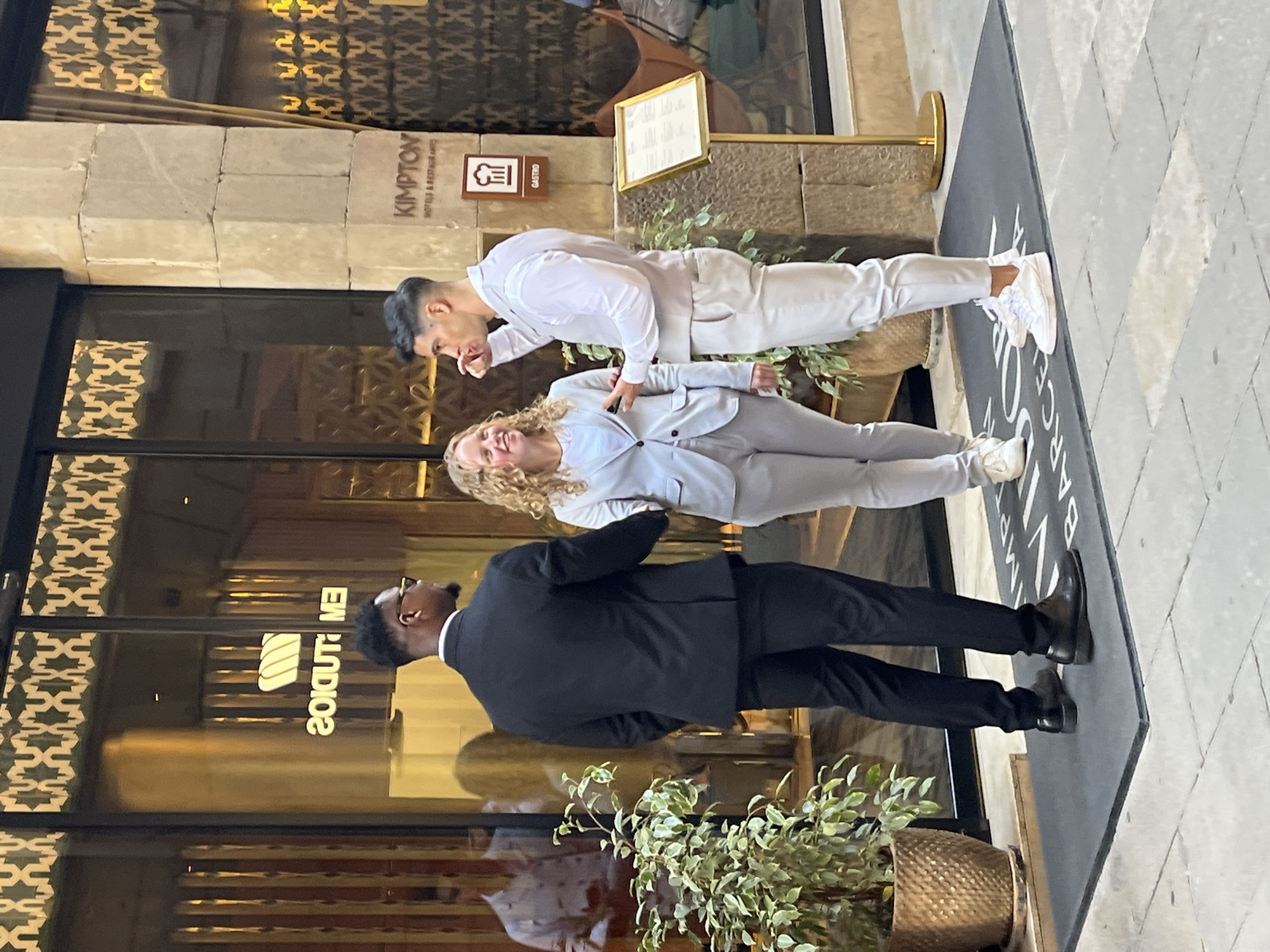 Three individuals are captured in a daytime, outdoor shot outside a modern building entrance in Barcelona, Spain. The scene appears to be the entrance of a hotel or restaurant, indicated by visible signage and architectural style.
On the left side of the frame, a man wearing a black suit, white shirt, and glasses stands with his back mostly to the camera, looking towards the building's entrance. In the center, a woman with curly blonde hair, dressed in a light grey suit and white sneakers, smiles pleasantly towards the viewer. To her right, a man with dark hair, wearing a white shirt, light grey vest, light grey trousers, and white sneakers, looks downwards and slightly away from the camera.
The background features a stone facade with the clearly visible text "KIMPTON HOTELS & RESTAURANT". Below this, a brown sign displays a fork and knife icon along with the word "GASTRO". The building's large glass windows feature intricate, decorative patterns. A large black doormat extends across the ground, showing partial text including "BARCEL" (likely part of "BARCELONA") and "ARCEL". A potted plant with green foliage sits on the ground near the building. Reflections on the glass windows also show reversed text, "2O!UDTUO", suggesting "STUDIO" or similar. The ground is paved with a light-colored, patterned tile. The overall impression is one of a professional or casual gathering in a stylish urban setting under clear, bright weather.