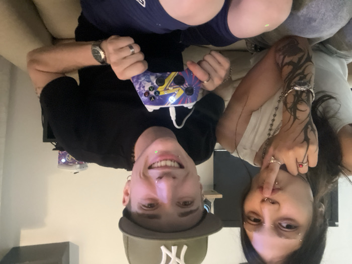 Two people are posing in an upside-down selfie. The person on the left is holding a game controller, wearing a black shirt, a New York Yankees hat, and a watch. They have a lip piercing and are smiling. The person on the right has dark hair, a tattoo on their arm, and several rings. They are holding a finger to their lips. They are also wearing a necklace. They both appear to be sitting on a beige couch inside a room with a television in the background.