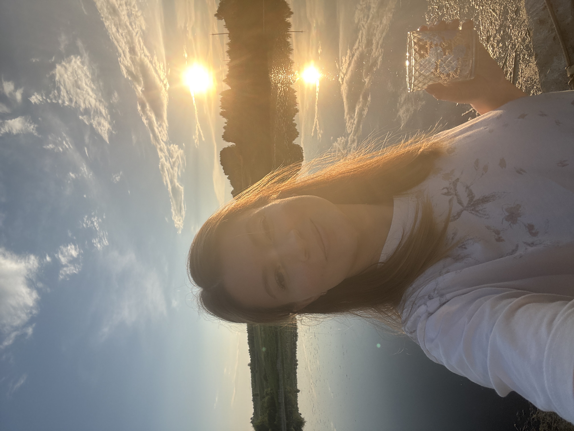 A young Caucasian woman is captured in a selfie outdoors, holding a decorative glass with a golden tint in her right hand. She is wearing a white t-shirt with a subtle pattern. The woman has long, reddish-brown hair that is illuminated by the setting sun, which is prominently featured in the upper right quadrant of the image. The sun casts a warm glow across the scene, reflecting on the calm surface of a body of water that occupies the middle ground. Behind the water, a line of trees forms the horizon, silhouetted against the vibrant sky. The sky itself is a blend of soft blues and oranges, with scattered white clouds catching the sunlight. The woman appears to be standing on the bank of the water, with dry, somewhat barren ground visible in the foreground. The overall atmosphere of the image is serene and tranquil, suggesting a moment of quiet enjoyment during sunset. The location is the city of Вассята, Russia.