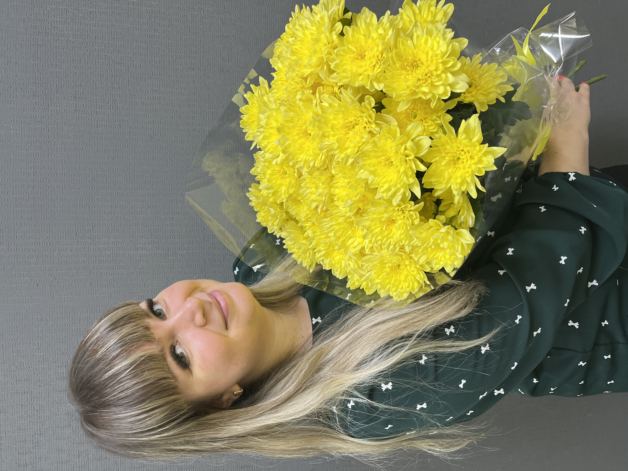 A woman is holding a bouquet of yellow chrysanthemums wrapped in clear plastic against a gray background. The woman has blonde hair with bangs, and she is wearing a dark green dress with white bow patterns. The scene is indoors. The location is not discernable from the image.