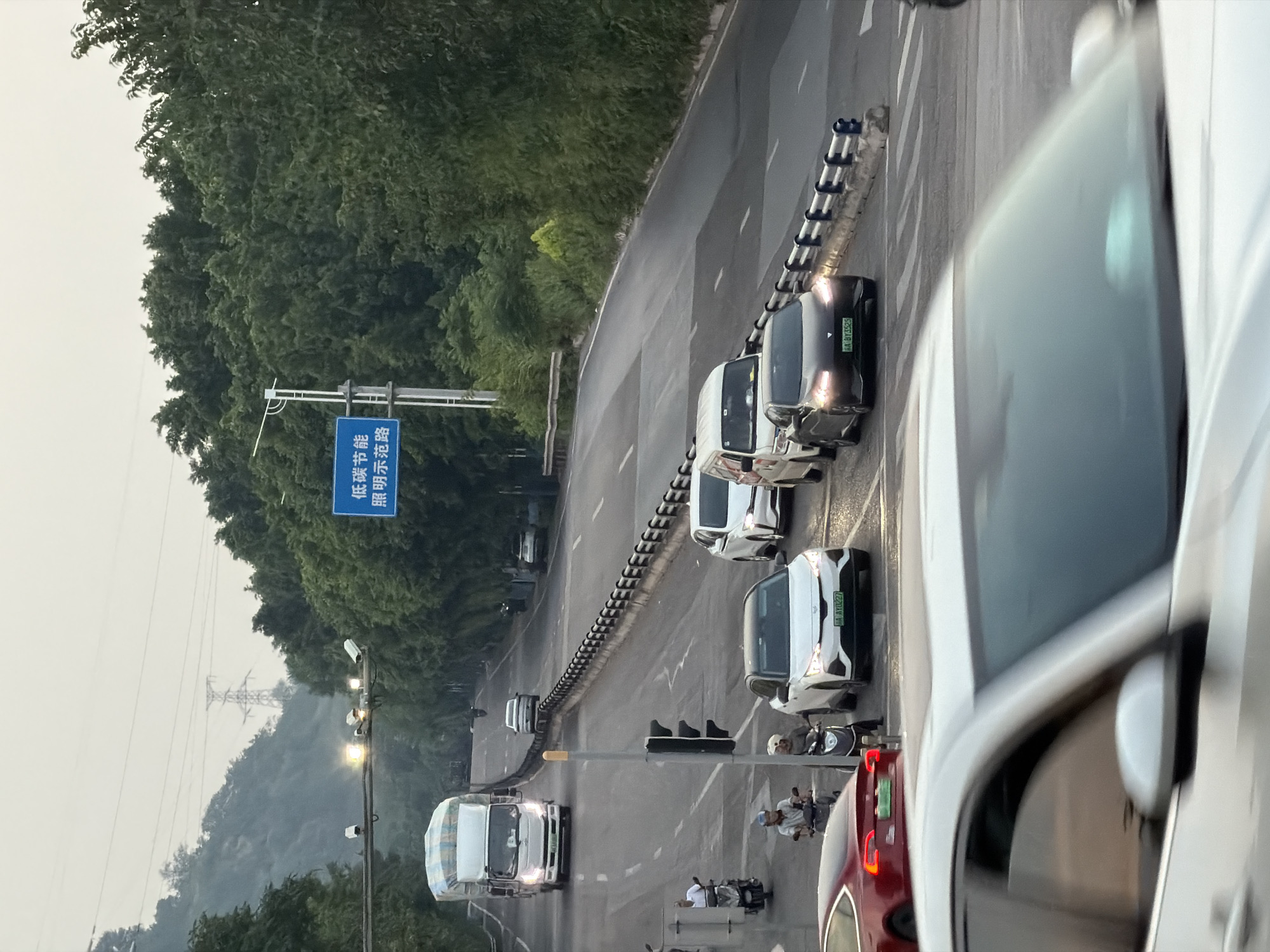 The image captures a road scene in Jiangjin District, Chongqing, China, likely during overcast daylight conditions. Multiple vehicles are present, including a large white truck with a covered load, several cars of various makes and colors, and a couple of motorcycles. Two individuals are seen on motorcycles, one in motion and the other stationary. A traffic light is visible, indicating potential traffic control. A blue directional sign with Chinese characters is posted on the right side of the road, reading "低碳节能 照明示范路" which translates to "Low-carbon energy-saving lighting demonstration road." The road is bordered by lush green trees and a metal barrier. Surveillance cameras are mounted on an overhead wire structure. The overall scene depicts a busy urban road with a focus on traffic and road infrastructure.
