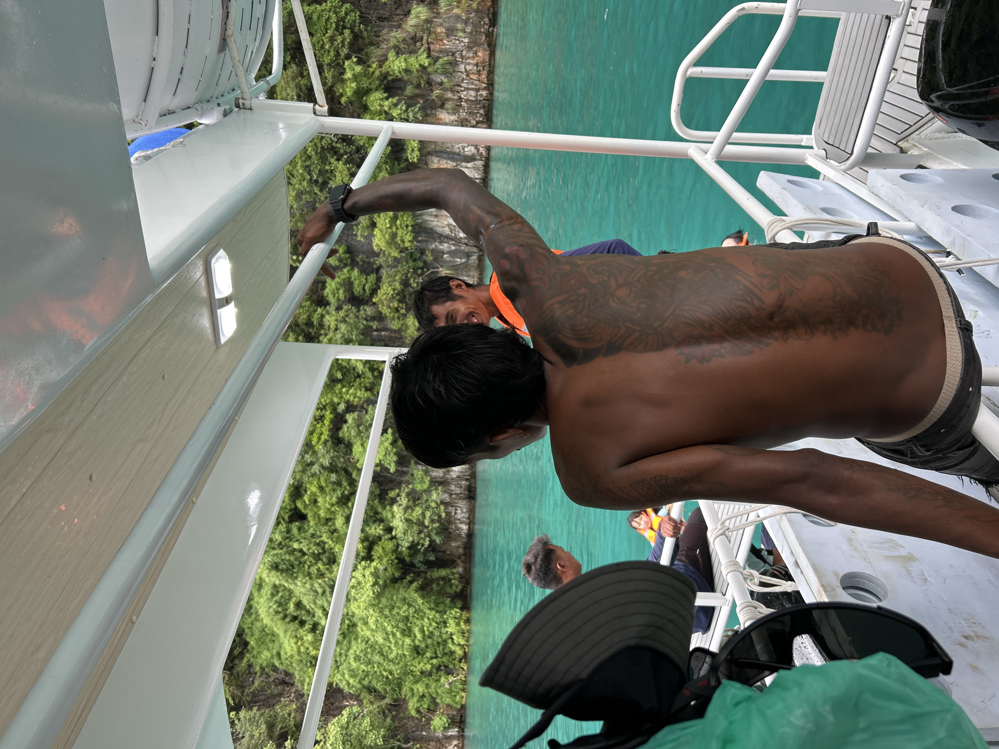In Ao Nang, Thailand, three individuals are aboard a boat on turquoise waters. The foremost person, with extensive back tattoos, faces away from the camera, their right arm raised. To their left, another man in an orange life vest smiles, looking toward the right. In the background, a child in a yellow life vest is visible. Lush green foliage covers the limestone cliffs that rise from the water, creating a scenic backdrop. The boat's white railing and deck are also prominent features of the scene.