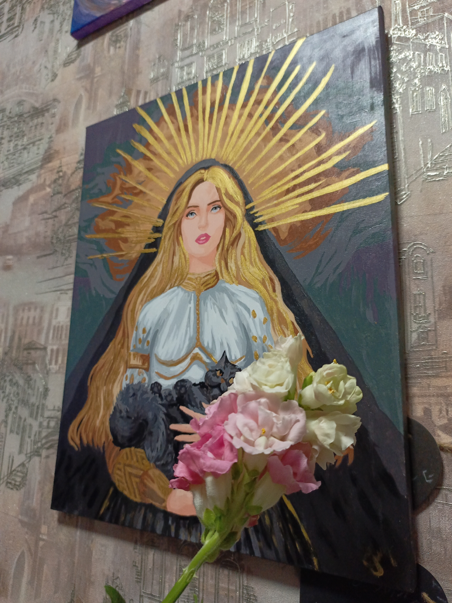 The image shows a painting depicting a woman with long blonde hair and a golden halo holding two black cats. She wears a white and gold garment, and a dark shawl drapes behind her. A bouquet of pink and white flowers is placed in front of the painting, partially obscuring the lower part of the artwork. The painting is hung on a wall with a patterned wallpaper. The location is Марковский, Russia.