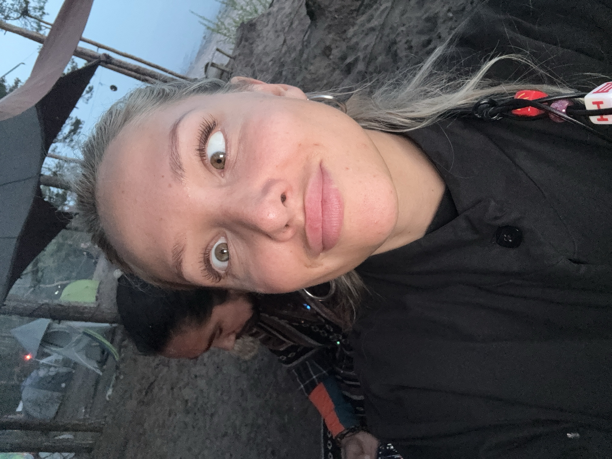 The image is a close-up selfie of a fair-skinned female with blonde hair and hazel eyes. She is wearing a black button-up shirt and hoop earrings. A person with dark hair and a patterned shirt is partially visible behind her. In the background, there are tents and trees, suggesting an outdoor camping setting. The location is likely Luzhki, Russia.