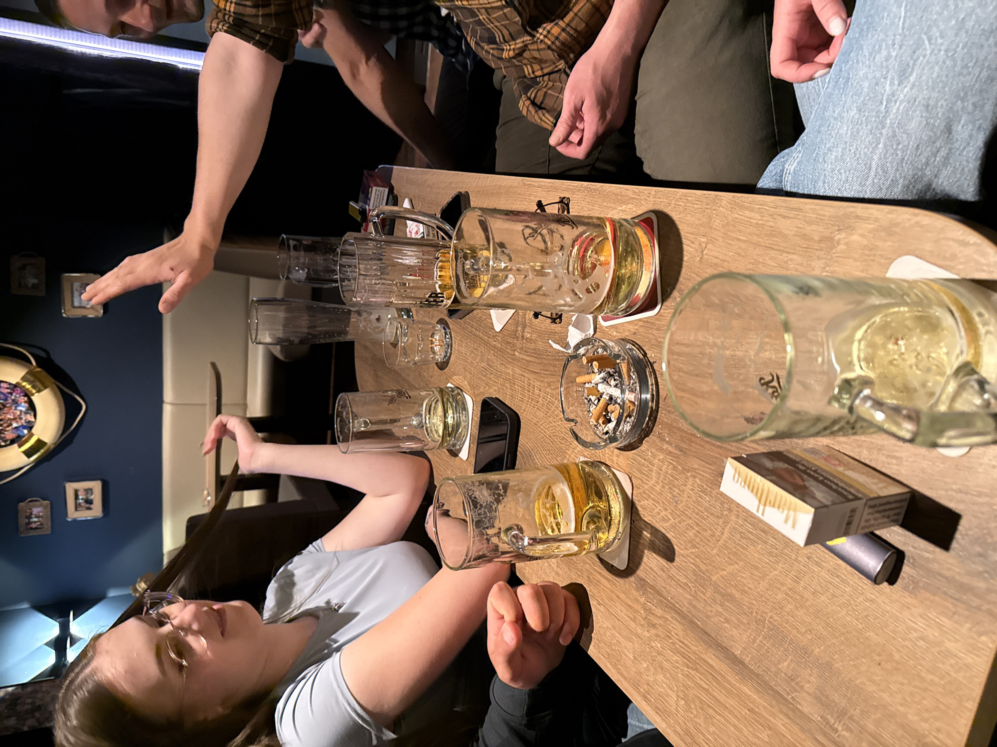 The image depicts a group of people gathered around a wooden table in what appears to be a dimly lit bar or pub. The setting has a nautical theme, with a life preserver visible on the wall and framed pictures.
In the foreground, a woman wearing glasses and a light blue shirt is seated on the left. She is holding a strand of her long brown hair. In front of her, on the table, are several beer mugs, an ashtray filled with cigarette butts, and a pack of Marlboro cigarettes next to a silver electronic device. One beer mug is partially filled, and another is nearly empty.
To the right of the woman, a person's arm is raised, with their hand extended as if in a gesture of stopping or greeting. This individual is wearing a plaid shirt. Further to the right, another person is seated, wearing a plaid shirt and dark pants. Their hands are resting on the table, with one hand showing a lighter.
In the background, a long, white booth seating arrangement is visible, along with additional tables and chairs. The overall atmosphere suggests a casual gathering. The presence of beer mugs, cigarettes, and an ashtray indicates that drinks and smoking are taking place. The overall scene is consistent with a bar or pub environment, potentially in Langeoog, Germany, given the query.