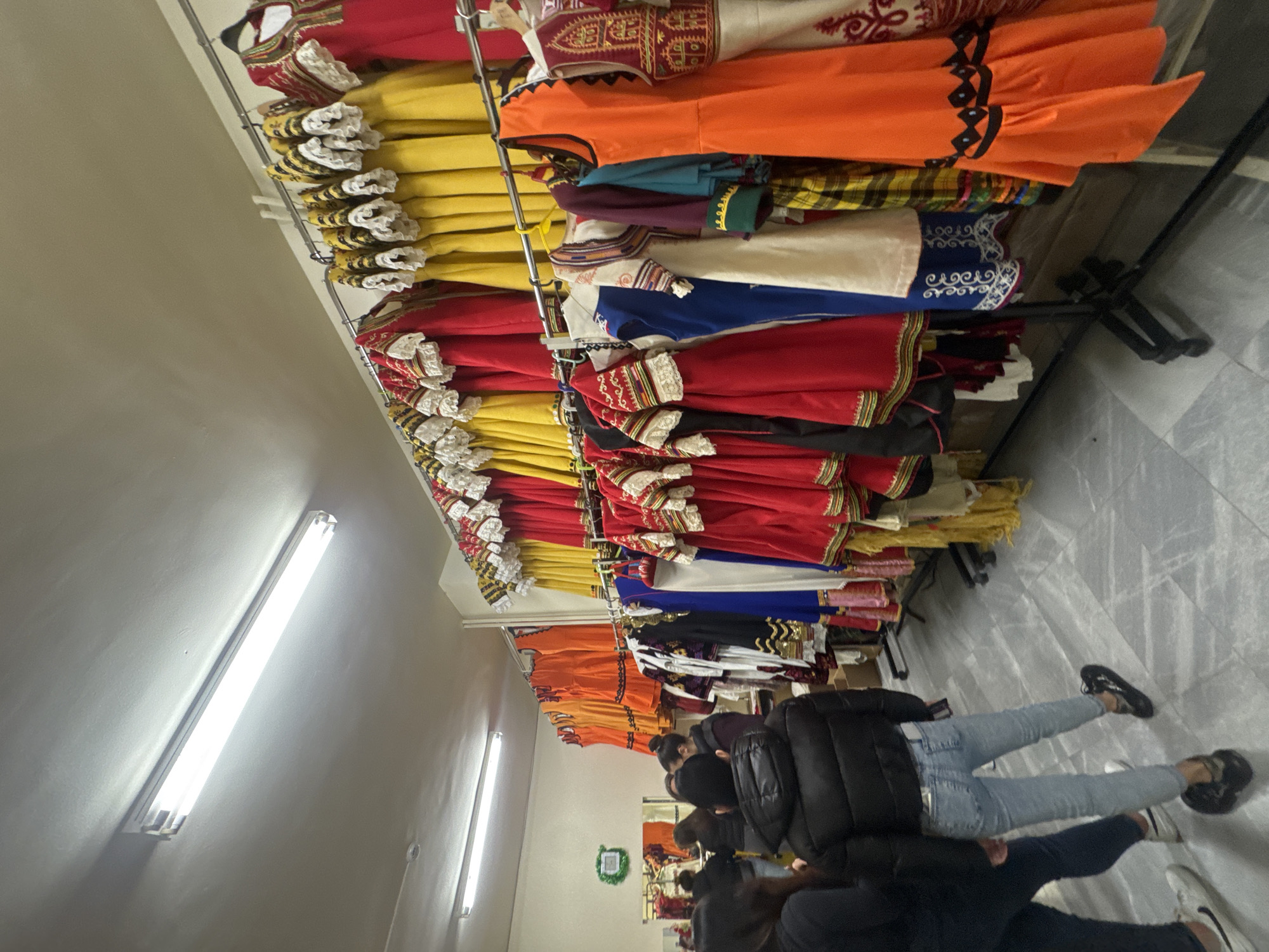 The image depicts an indoor scene, likely a costume shop, storage area, or backstage preparation room, filled with a vibrant array of traditional folk costumes in Sofia, Bulgaria. The garments are predominantly in bright red, yellow, and orange, meticulously organized on metal clothing racks that extend across the length of the room.
Many of the costumes feature elaborate detailing, including intricate embroidery on the bodices and sleeves, often accented with gold threads, as well as ruffled white lace trim, particularly noticeable on the yellow and red dresses. Other costumes in blue, white, and various plaid or patterned fabrics are also visible, showcasing a rich variety of traditional attire.
In the lower portion of the image, several people are seen, mostly from behind, appearing to browse or select costumes. One individual is prominent in light blue jeans, a dark puffer jacket, and dark sneakers. The other people are dressed in casual, dark clothing, contrasting with the colorful traditional garments. Their heads are generally bowed, suggesting they are inspecting the hanging items closely.
The setting features a simple interior with a grey tiled floor and plain light-colored walls. The space is well-lit by several long, bright fluorescent lights mounted on the ceiling. A small, circular green wreath is visible on a distant wall, adding a subtle decorative touch. There is no visible text in the image, and the expressions of the individuals are not discernible. The scene suggests an active environment centered around the preparation or selection of traditional clothing.