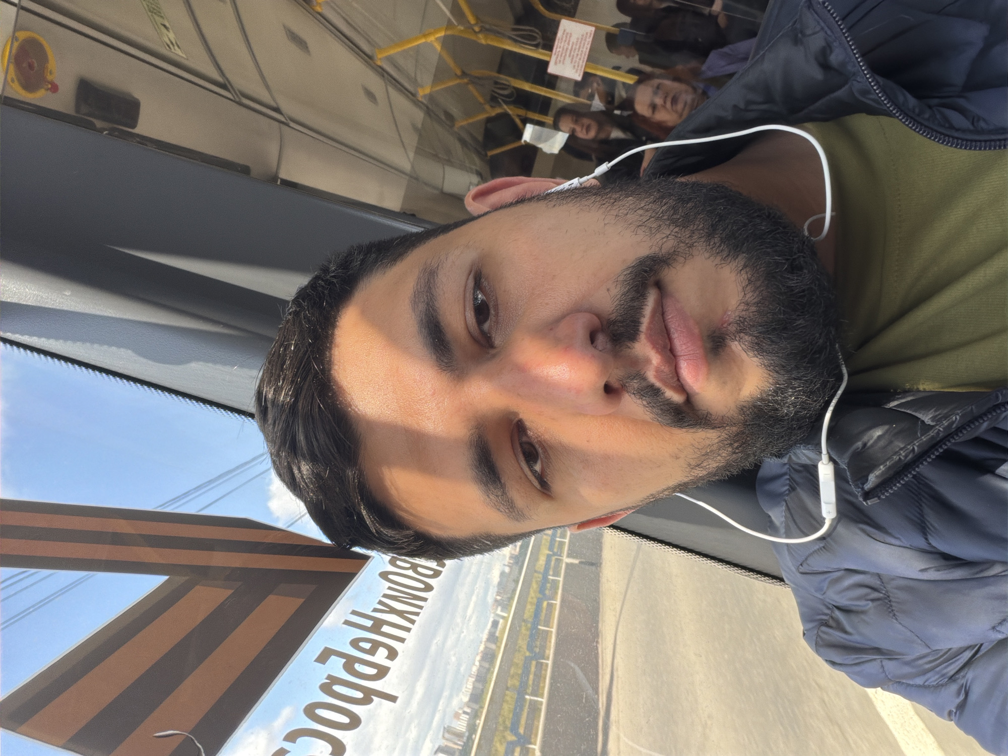 A medium indoor shot shows a man in a bus or train, turned to the left. He has short dark hair, brown eyes, brown skin, and a black beard. He is wearing white earphones and a dark-blue puffy jacket over a green shirt.
Outside the window is a clear blue sky, and a view of what appears to be the city of Krasnoyarsk, Russia. The image is rotated 90 degrees counter-clockwise. In the background, other passengers are visible. A sticker is on the bus window with a letter “V” on it.