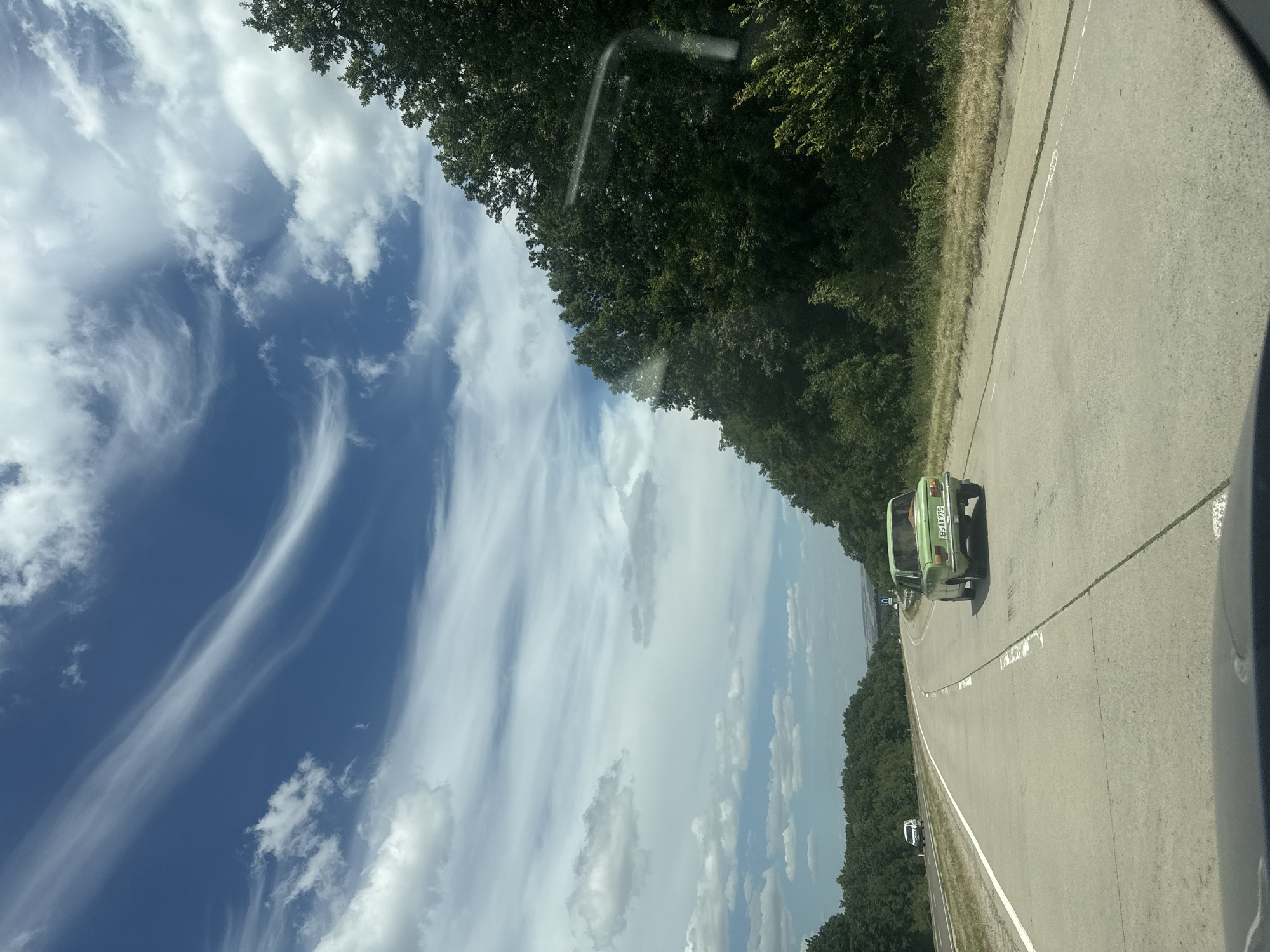 The image depicts a rural highway scene with a clear blue sky adorned with wispy white clouds. The road, paved with concrete, curves to the right, bordered by dense green trees on the right side and a grassy shoulder on the left. A light green, older model car is traveling ahead on the road, its license plate reading "BS AA 775". In the distance, a white SUV is visible on the highway, and farther down the road, a sign with a blue 'U' shape can be seen. The foreground shows the dashboard of a vehicle, suggesting the photo was taken from inside a car. The location is the road leading to or from the city of Râzeni, Moldova.