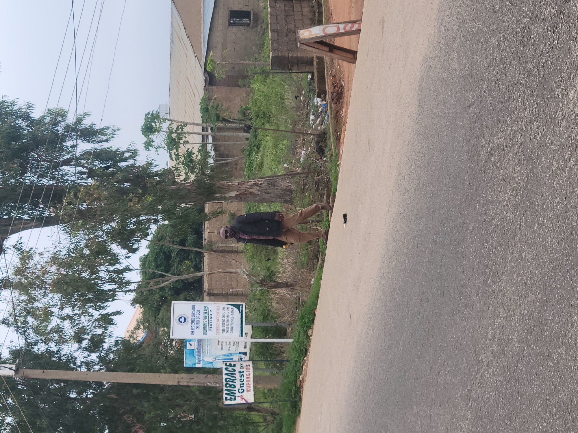 The image depicts a street scene in Jos, Nigeria. In the foreground, a paved road stretches across the frame. On the left side of the road, a man in a dark jacket and tan pants is walking away from the camera. He is wearing sunglasses and appears to be holding a yellow object in his right hand. Behind him, several signs are visible, including one for "EMBRACE GUEST INN KUFANG JOS" and another with the logo of "THE REDEEMED CHRISTIAN CHURCH OF GOD SOLOMON'S PORCH AREA PLATEAU 2". The background shows a mix of vegetation, trees, and buildings with corrugated metal roofs. To the right of the road, a-frame sign stands partially visible. The overall environment suggests an urban or semi-urban setting.