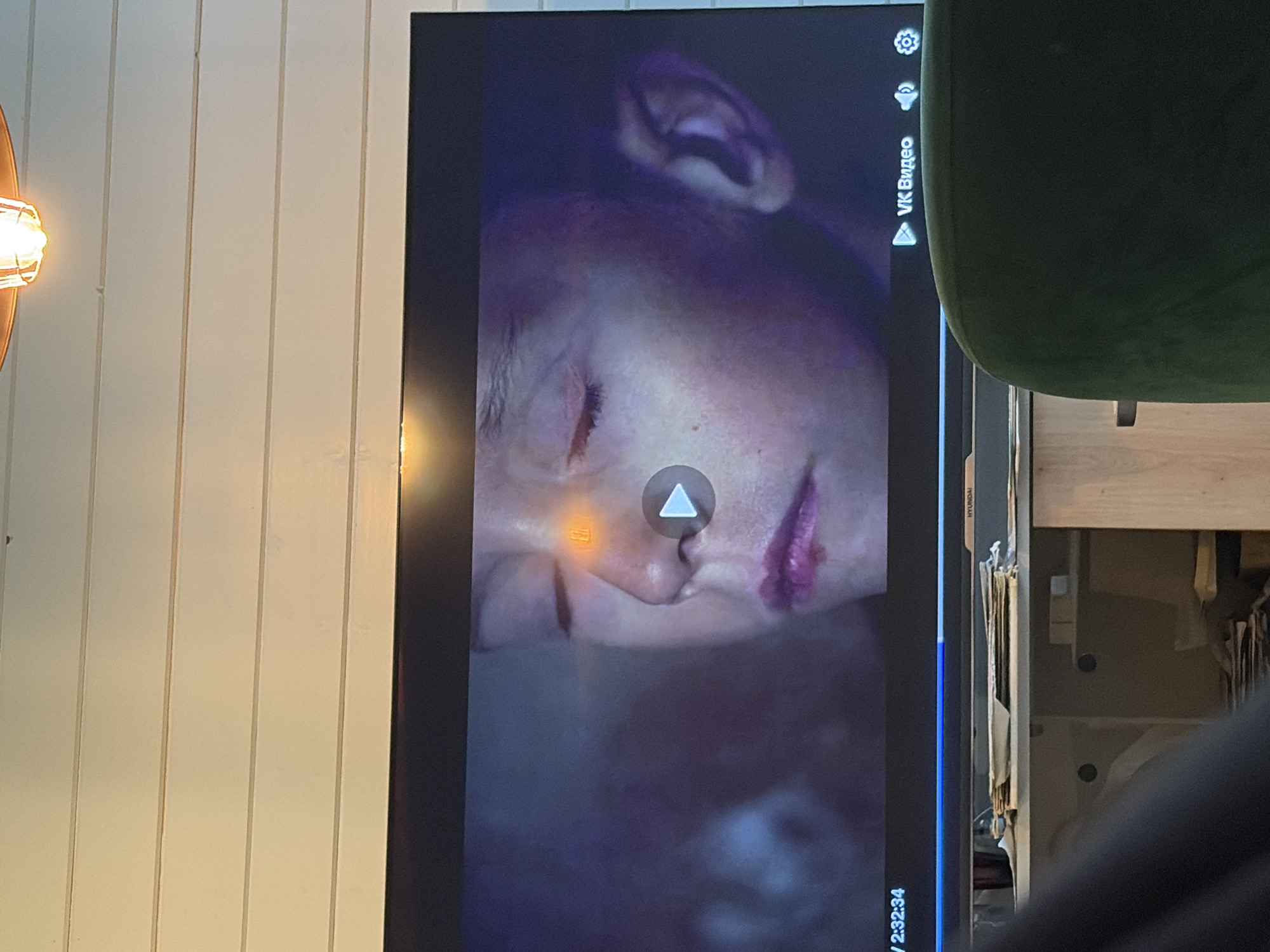 The image shows a close-up view of a television screen displaying a scene with a young boy with his eyes closed. An orange glow emanates from his forehead, and he has bright pink lips. A play button is visible on the screen, along with text that reads "VK Видео" (VK Video). In the bottom left corner, a timestamp "2:32:34" is displayed. The television is positioned above a piece of furniture, possibly a cabinet, on which a stack of papers and other items are placed. In the lower right portion of the image, a dark green chair is partially visible. The background above the television consists of horizontally aligned wooden planks. The scene appears to be an indoor setting. The location of Snegiryovka, Russia, is not discernible from the image itself.