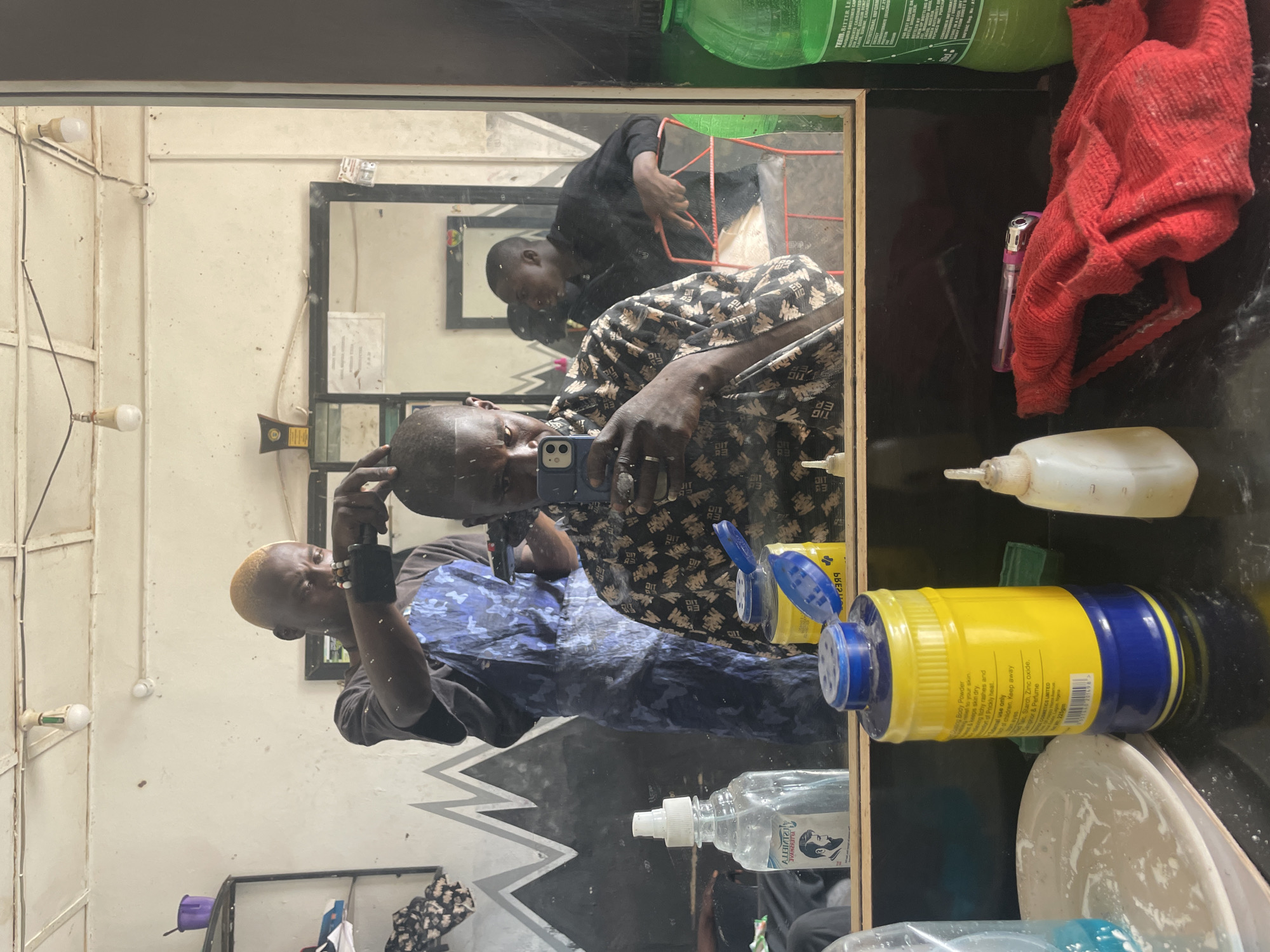 The image shows a barbershop setting in Durimi, Nigeria, reflected in a mirror. Two men are visible: one is taking a selfie with his phone, wearing a patterned shirt, and the other is behind him, possibly cutting hair. The barbershop has simple, functional decor, with exposed light bulbs on the walls. Various hair care products are placed on a surface in front of the mirror, including a yellow and blue container of body powder. Other items include a red cloth, a lighter, a bottle with a nozzle, and another container.