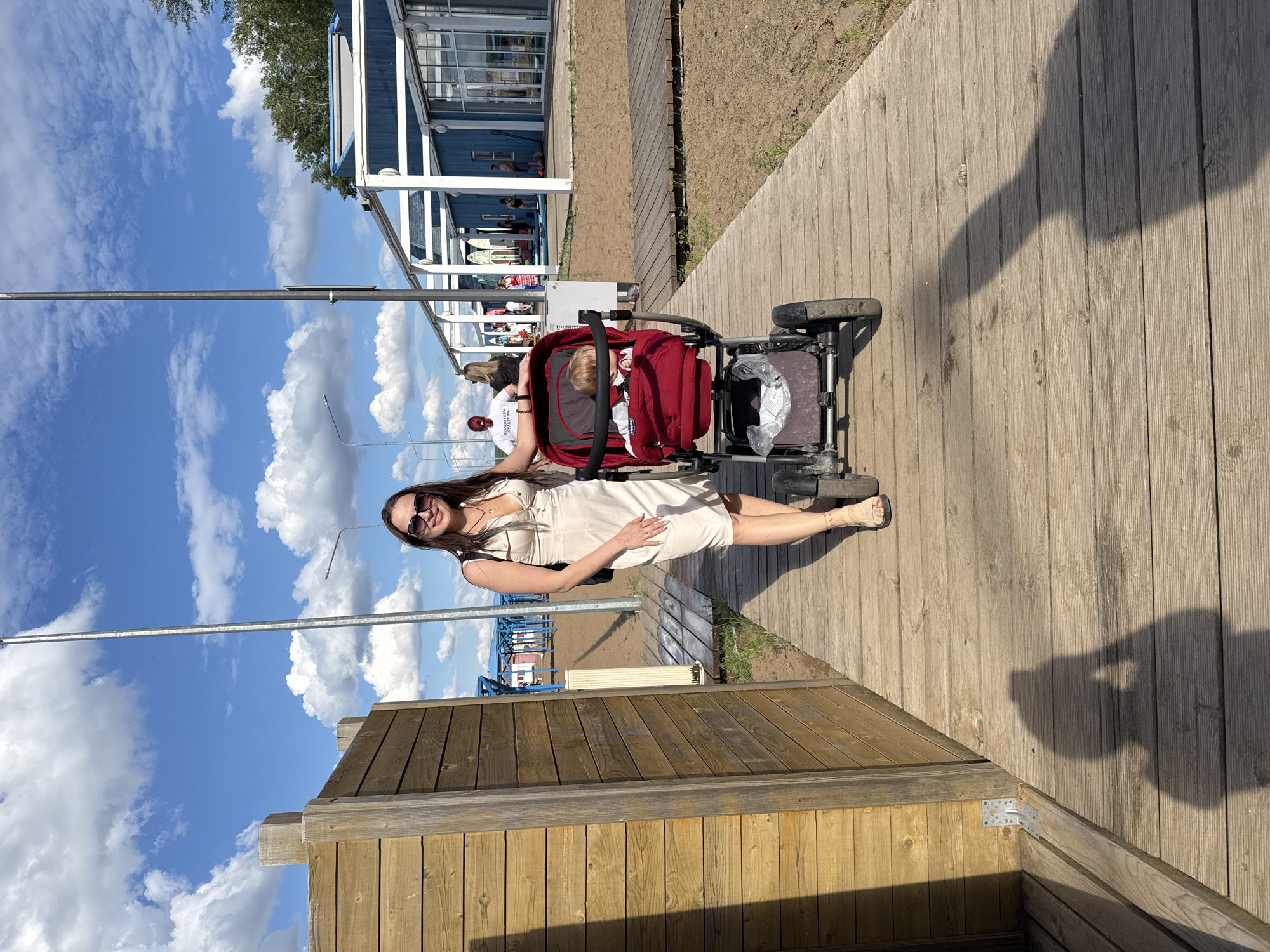 A woman wearing a light-colored dress and sunglasses is posing next to a red stroller. The stroller contains a child. They are on a wooden boardwalk, which is a part of a beachside recreational area. Behind them, there is a blue building with white trim, possibly a cafe or changing room, with people visible inside and around it. The background also shows a sandy beach and the sky with scattered white clouds. The sun is casting a shadow on the boardwalk, suggesting it is daytime, likely late afternoon due to the warm lighting. The scene appears to be a casual outing, possibly at a beach resort or park in Chaikovsky, Russia. There is no visible text in the image.