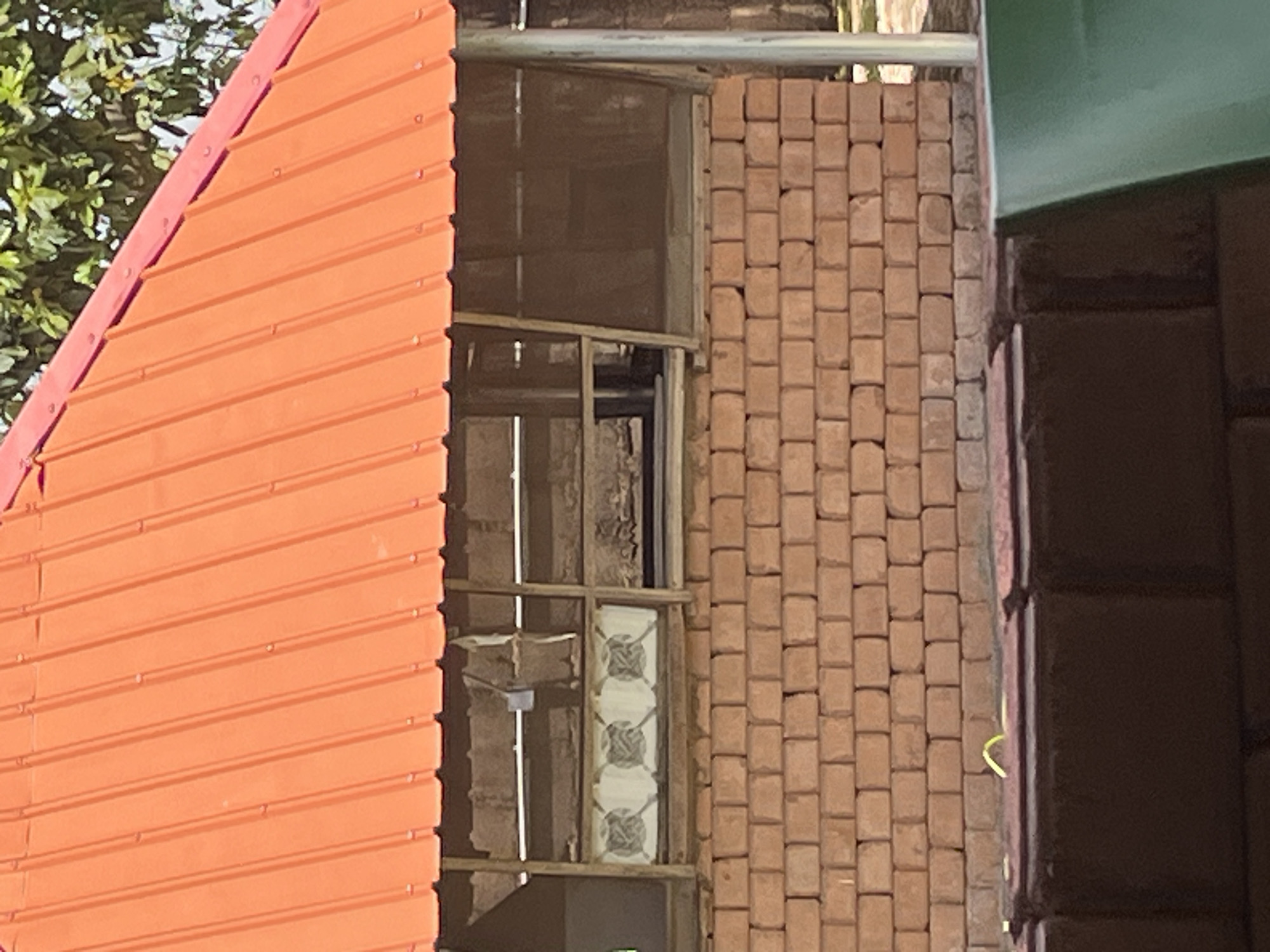 This media file depicts an outdoor scene featuring parts of a building under construction. On the left, an orange corrugated metal roof slopes upwards, suggesting an angled structure, possibly a shed or a section of a house. To the right of the roof, a wall constructed from small, reddish-brown bricks is visible. The brickwork is detailed, showing individual bricks laid in a staggered pattern.
In the center of the image, there is an open window frame. The frame appears to be made of wood and is partially obscured by the brick wall. Inside the window, there are glimpses of wooden structural elements and what looks like a piece of fabric with a repeating geometric pattern, possibly a decorative panel or a piece of cloth. The window itself is dark, suggesting it may be covered by a screen or is simply too deep to illuminate. Above the window, a horizontal metal pipe or beam runs across the frame.
To the far right, a dark brown, possibly wooden, wall or panel is visible. Above this section, a curved, green surface is present, which could be part of a large water tank or another structure. A thin, yellow, curved object is visible near the edge of the brown panel, its purpose unclear. The lighting suggests it is daytime, with sunlight illuminating the scene and casting subtle shadows. The overall impression is one of ongoing construction or renovation in an outdoor setting. No people are visible.