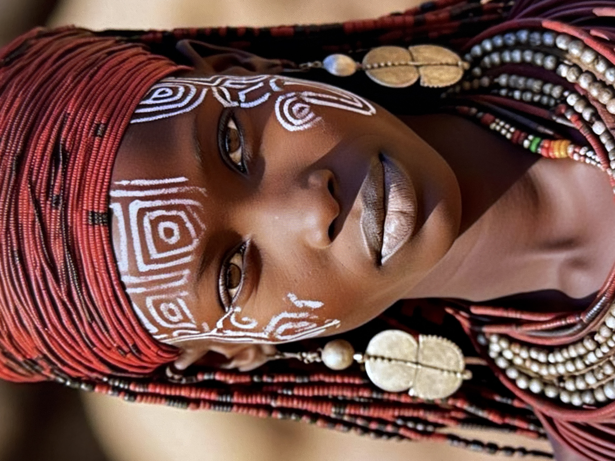 A close-up portrait shows a young person with dark skin and intricate white face paint. They are wearing a red headdress, large dangling earrings, and layered necklaces made of beads and red cords. The background is a blurred, neutral tone, suggesting an outdoor, possibly arid environment. The image does not depict any location in Orihuela, Spain.