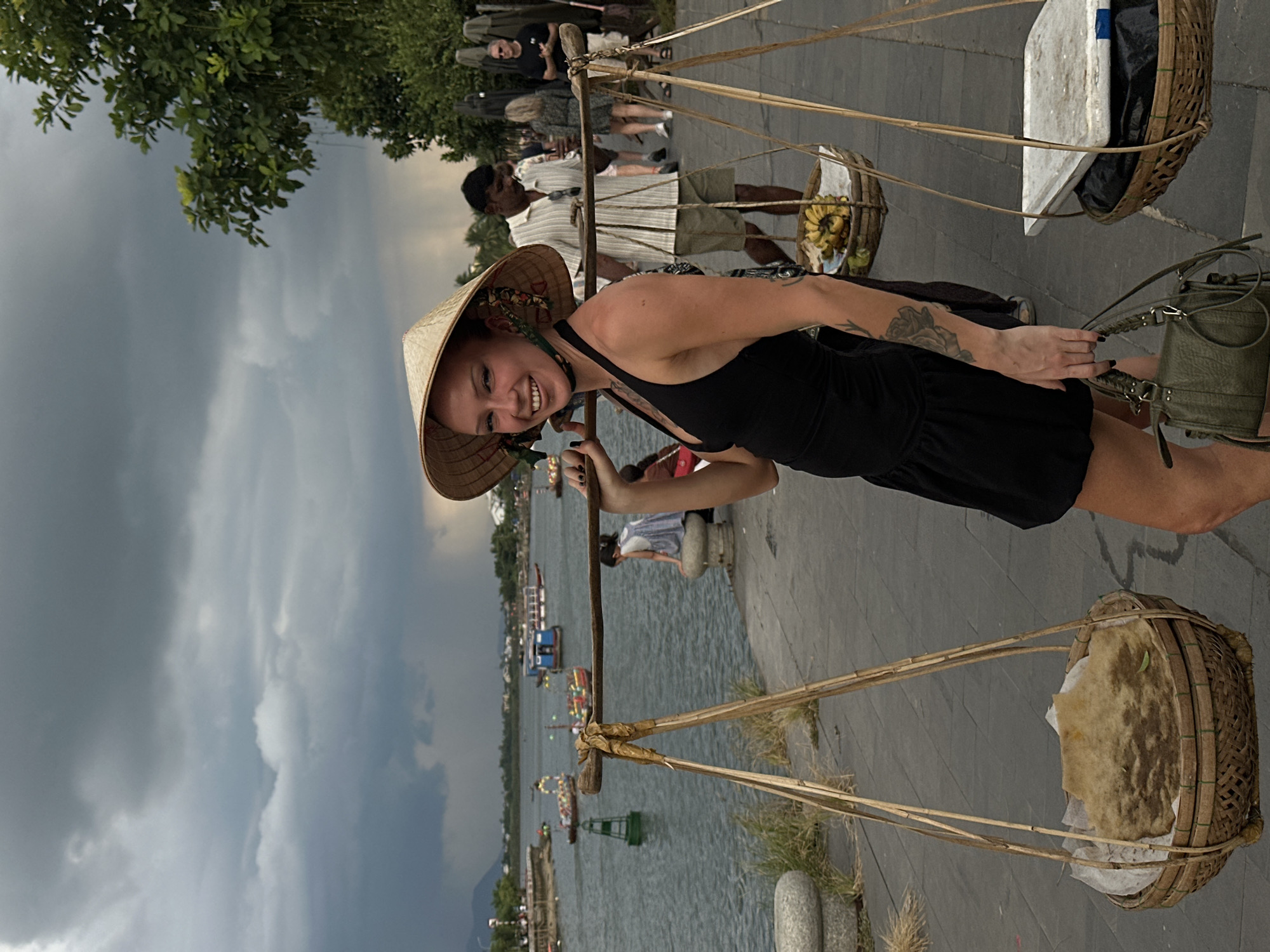 In Hoi An, Vietnam, a woman with a wide smile wears a conical hat and a black halter dress, carrying a traditional shoulder pole laden with two woven baskets. The left basket contains a large, flat, round food item, possibly bread or a cake, wrapped in paper. The right basket holds a bunch of bananas and what appears to be a white board. In the background, a river flows, with several boats docked or moving on its surface. People stroll along the paved riverwalk, and lush greenery and trees frame the scene. The sky is overcast with dramatic, dark clouds.