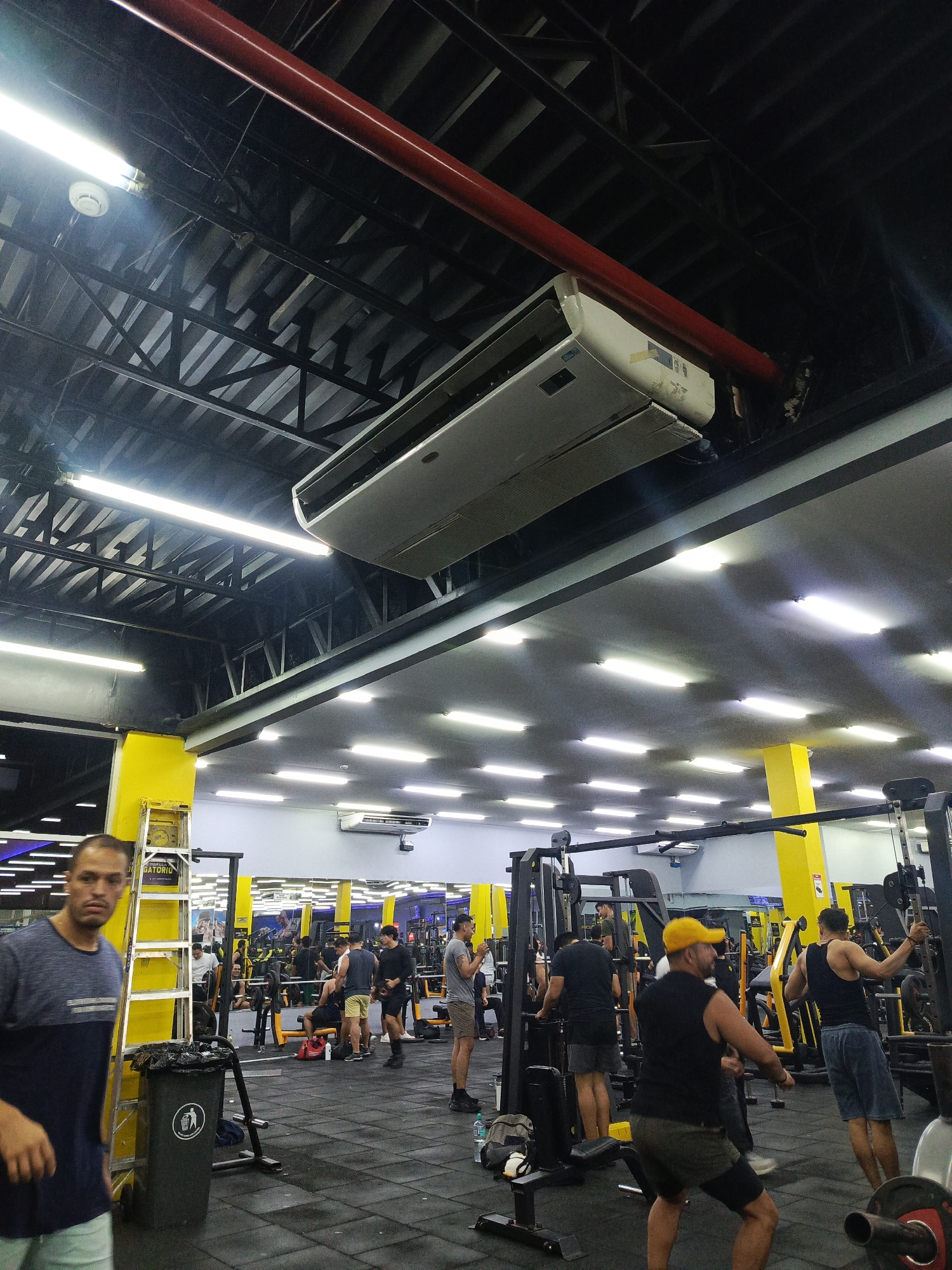 The image captures an indoor scene within a brightly lit gymnasium, likely located in Guayaquil, Ecuador, as suggested by the presence of yellow structural elements and the overall aesthetic common in such establishments in the region. The ceiling features exposed black metal beams and is adorned with numerous fluorescent light fixtures and two prominent air conditioning units. A large red pipe runs horizontally across the upper portion of the frame, positioned above one of the air conditioners.
The gym floor is covered in black rubber mats, and various exercise machines, including weight racks and benches, are visible. Several individuals are engaged in different fitness activities. In the foreground on the left, a man wearing a blue and grey t-shirt and light-colored shorts is partially visible, looking towards the camera with a neutral expression. To his right, a tall ladder with a yellow crate on it leans against a bright yellow pillar. A dark gray trash bin is situated next to the ladder.
Further into the gym, a group of men are working out. One man in the mid-ground is wearing a yellow cap and a black sleeveless shirt, performing an exercise. Another man, on the right side of the frame, is wearing a black tank top and blue shorts, appearing to be engaged with gym equipment. Other gym-goers are scattered throughout the space, with some lifting weights and others standing or walking. Mirrors line the back wall, reflecting the gym equipment and people, creating a sense of depth. The overall atmosphere appears to be one of active exercise and a typical gym environment.