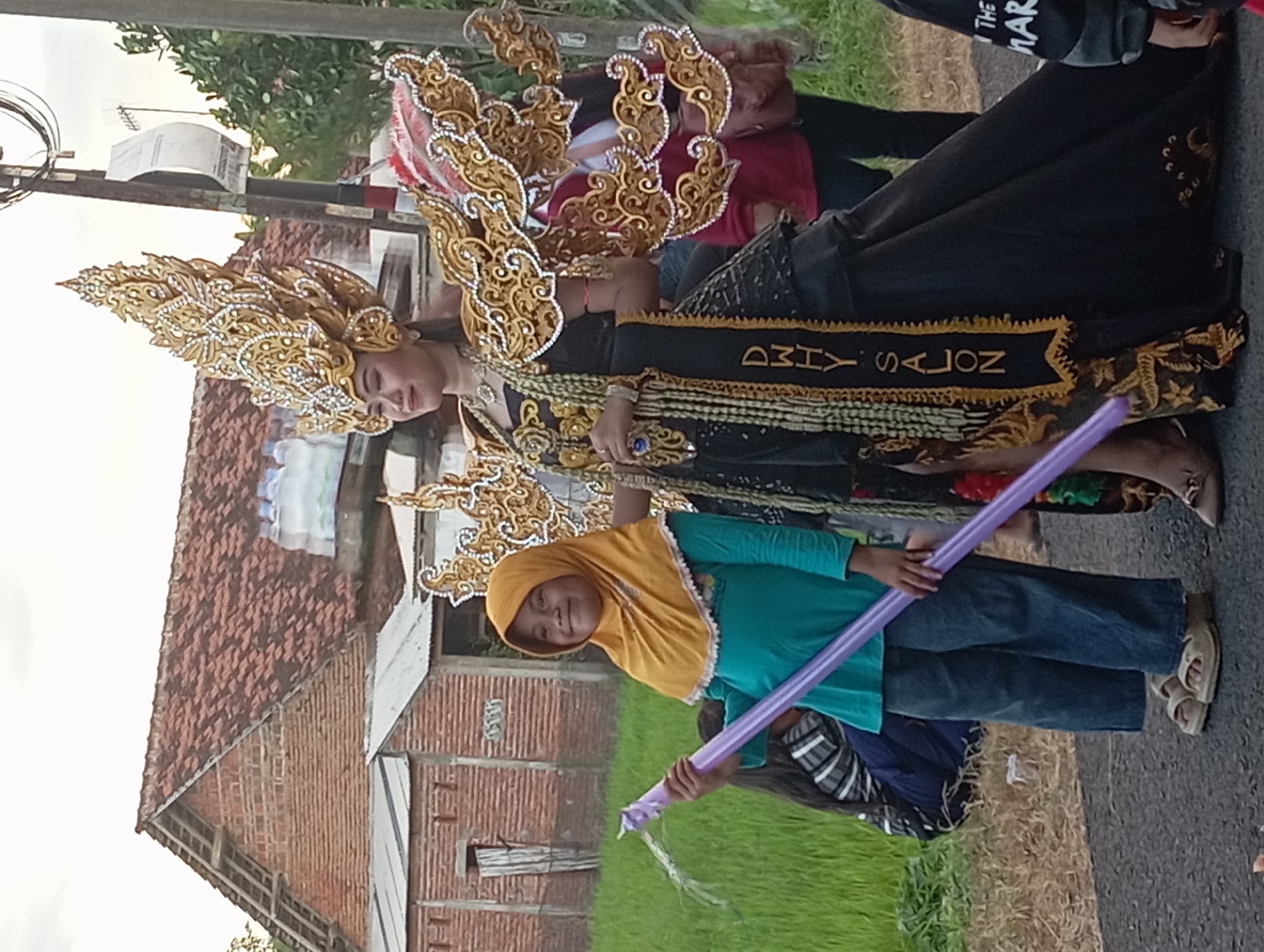 In Waung, Indonesia, an individual in an elaborate, gold-colored headpiece and ornate black and gold garment is walking on a paved road. The garment has the words "DMY SALON" on it. Standing beside them is a child with a yellow hijab, a blue shirt, and jeans, holding a long purple balloon. They are on a road next to a field of green grass and a brick building. Overhead are electrical wires.