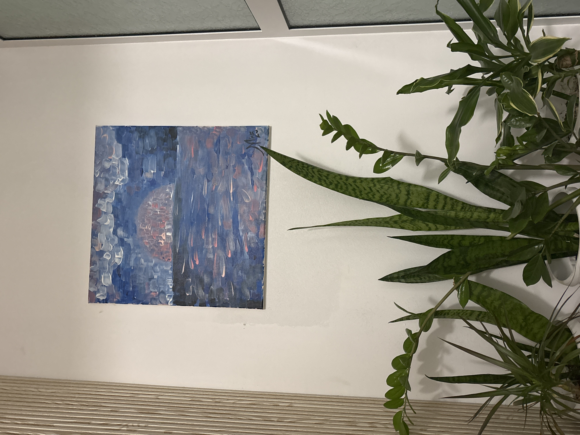 The image shows an interior setting with a white wall on which a square abstract painting hangs. The painting features shades of blue and gray with hints of red, depicting a somewhat nebulous scene. To the right of the painting, a collection of green plants fills the frame, including what appears to be a snake plant and another vining plant with small round leaves. Beneath the wall, there is a surface with a striped pattern. Above the wall, a horizontal window frame is visible. The image does not contain any information about people, their actions, interactions, or a city location.