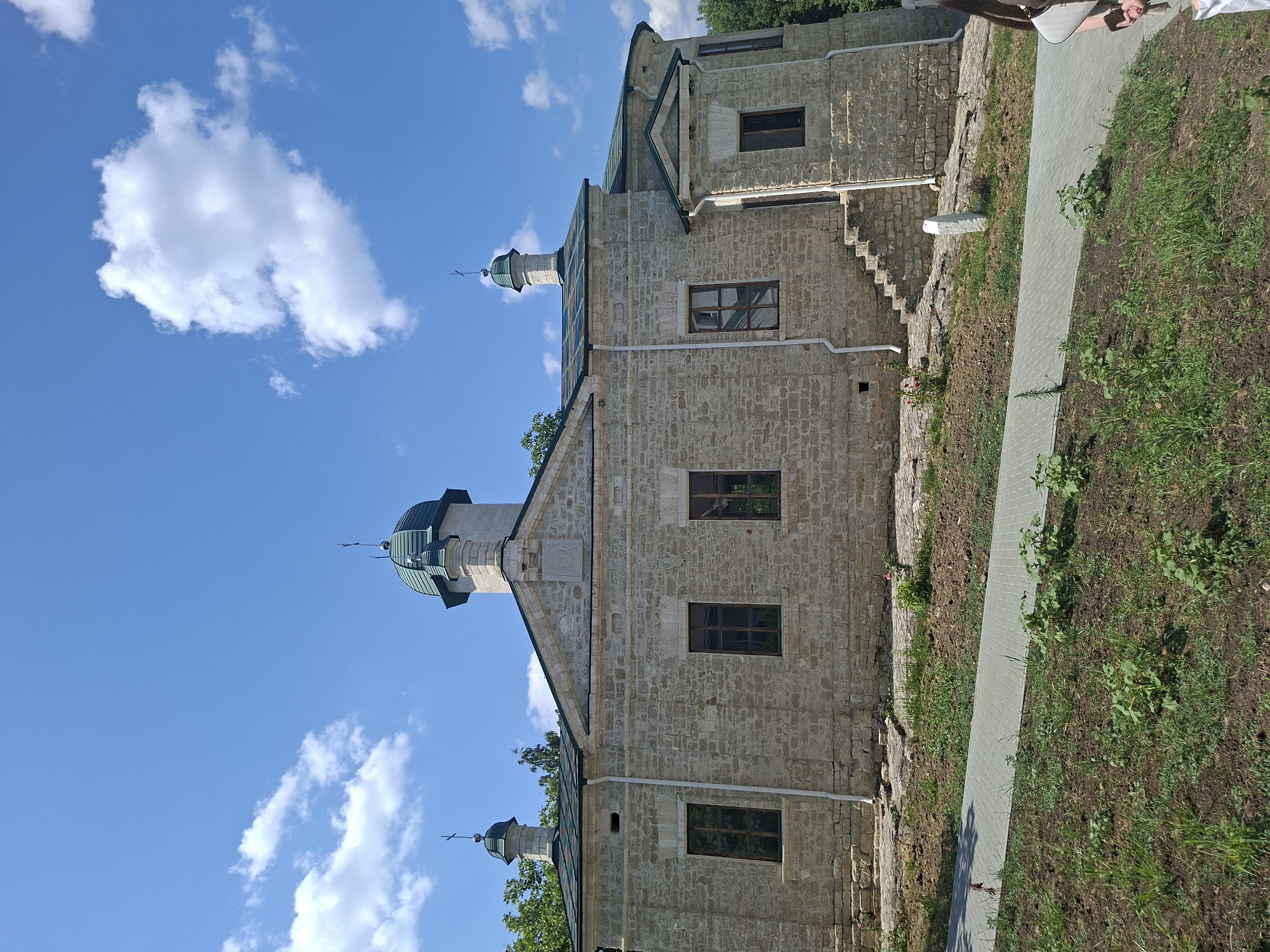 The image depicts a stone building with a green domed roof and two smaller green domed structures topped with crosses, suggestive of a church or monastery. The building's facade is constructed from rough-hewn light-colored stones, with several windows. A set of stone stairs leads up to an entrance on the right side of the building. A paved walkway is visible in the foreground, bordered by grassy areas and soil with some small plants. In the background, trees are visible. The sky is blue with scattered white clouds. The scene appears to be outdoors on a sunny day. The building is identified as the Condrița Monastery in Condrița, Moldova. A portion of a person with dark hair is visible on the far right edge of the image.