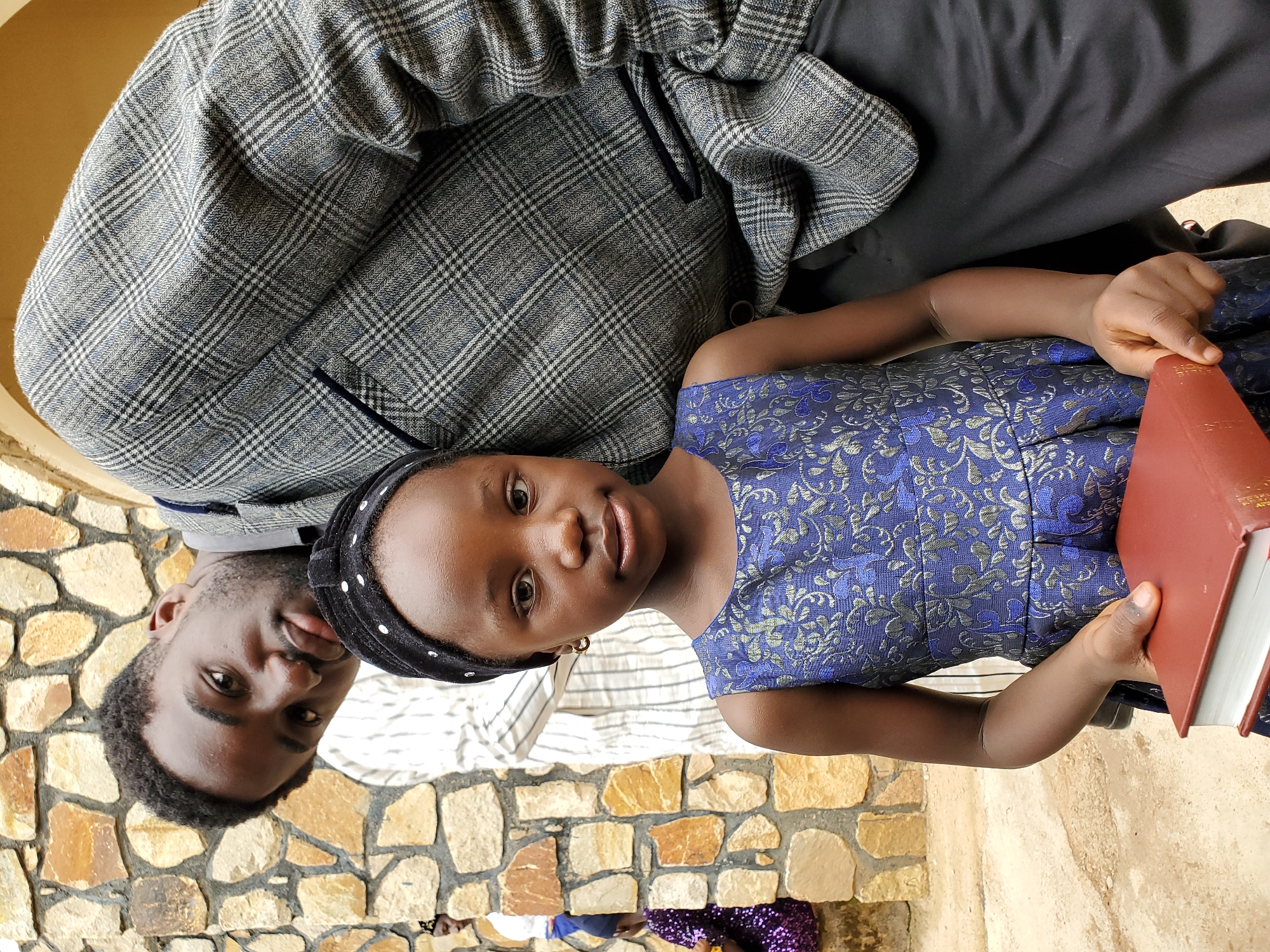 A medium shot shows a young girl and a man looking at the camera. The girl is wearing a blue patterned dress and a black headband with white polka dots and is holding a red book. The man has short black hair and is wearing a gray plaid jacket. They are standing in front of a wall made of tan-colored stones. The location is in Jos, Nigeria.