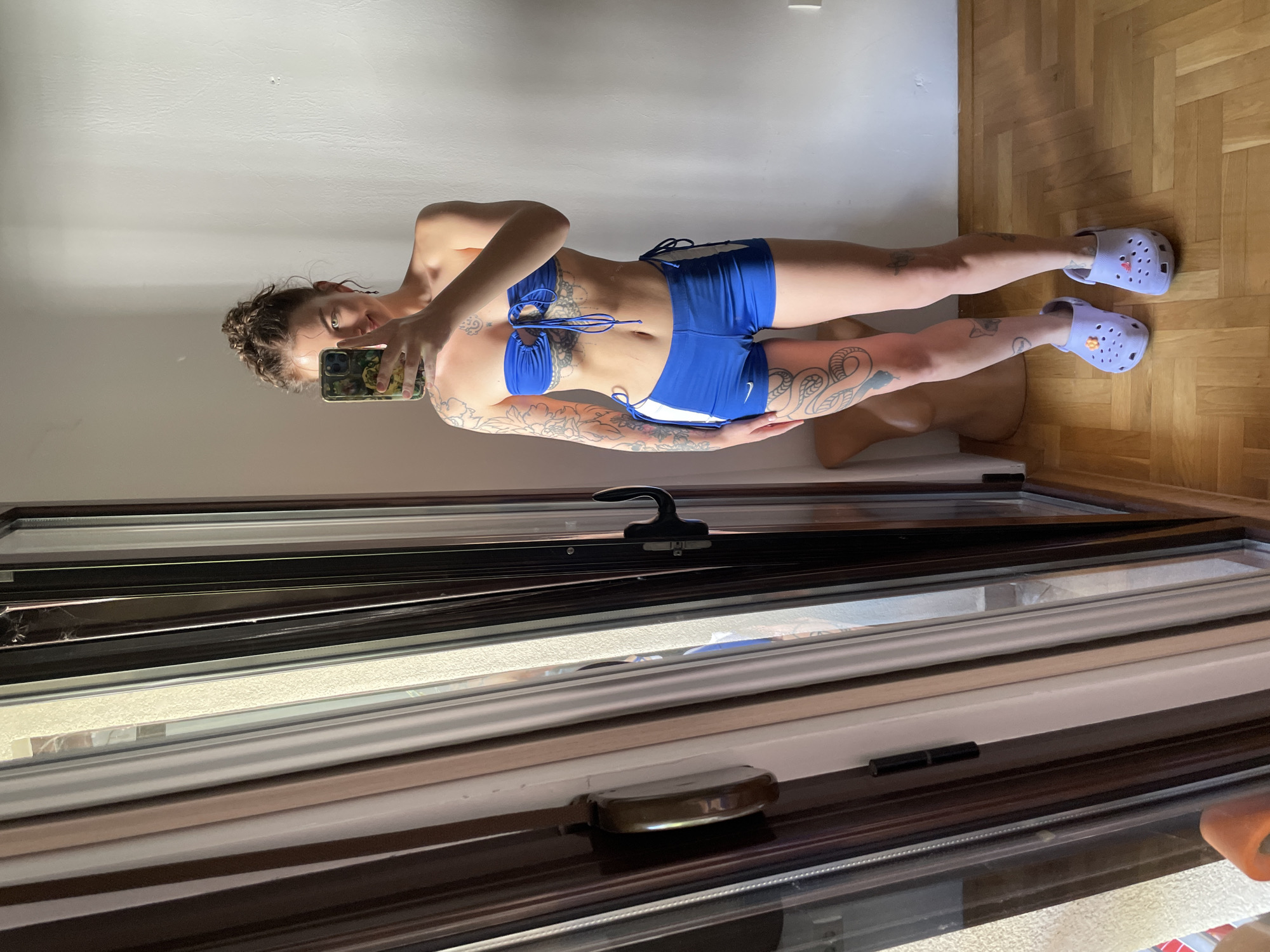 The image captures a full-body mirror selfie of a young adult female within a residential room in Boreti, Montenegro. The subject stands centrally, slightly angled, holding a dark smartphone with a multi-colored, possibly camouflage-patterned case, obscuring the upper portion of her face; her right eye and a partial smile are visible. Her light brown, curly hair is styled in a messy bun, and she wears a small dark earring on her right ear.
She is dressed in a two-piece athletic set consisting of a strapless royal blue bandeau top with central ruching and criss-cross blue string ties, and matching royal blue athletic shorts featuring white vertical stripes on the outer thighs and side drawstrings. A Nike logo is visible on the left thigh of the shorts. On her feet, she wears light purple Crocs-style clogs, with an orange floral charm on the left shoe and a pink heart charm on the right.
Extensive tattoos adorn her body: intricate floral and leafy patterns cover her left shoulder and arm, fine-line ornamental designs are visible on her abdomen and ribcage, a large serpentine tattoo is prominent on her left thigh, and smaller, less defined tattoos are on her right calf.
To the bottom left, partially visible behind her left leg, is a light brown, headless, armless female mannequin torso leaning against the wall.
The room's background features a textured white wall. The floor is light-colored wooden parquet laid in a herringbone pattern. On the left side of the frame, a multi-panel sliding glass door or window system is visible, characterized by dark brown/bronze and lighter brown/beige frames. Various dark-colored handles and hinges are observable on the window frames. Natural light enters from the left, casting illumination across the scene.