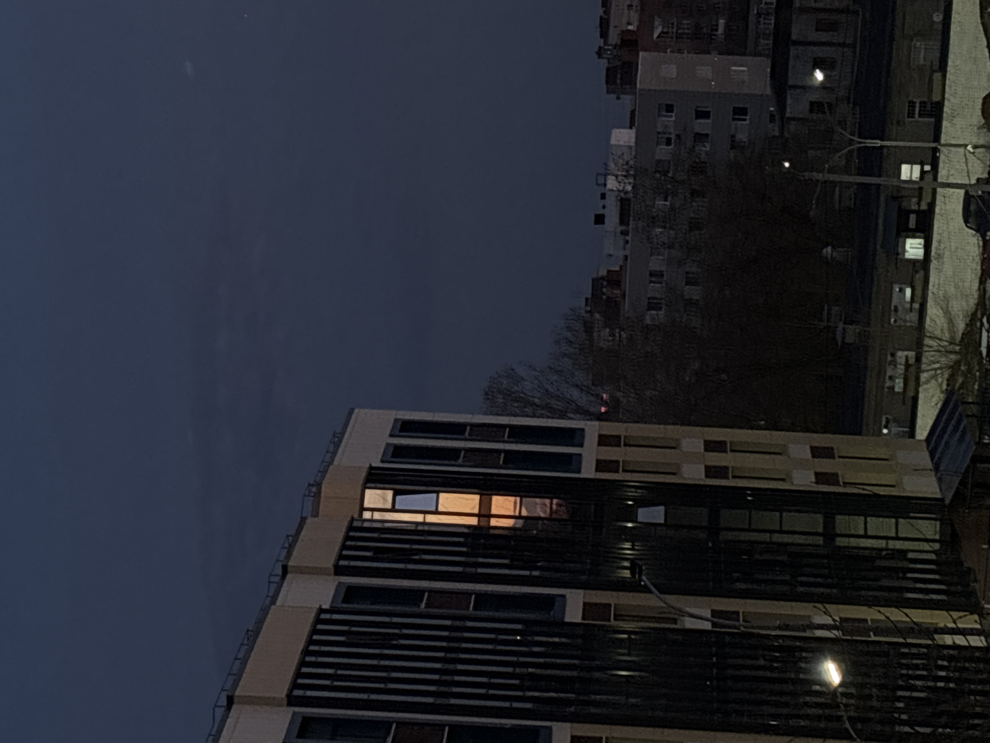 A low-angle shot presents a night cityscape in Perm, Russia. A modern high-rise building dominates the lower portion of the frame, its facade featuring linear patterns, illuminated by an internal orange glow emanating from one of its windows. Streetlights are visible below. In the distance, another residential building complex is seen, punctuated by intermittent lights from various apartments. The sky above is a dark, deep blue. Bare tree branches are visible in front of the taller building. A street lamp is positioned in the foreground.