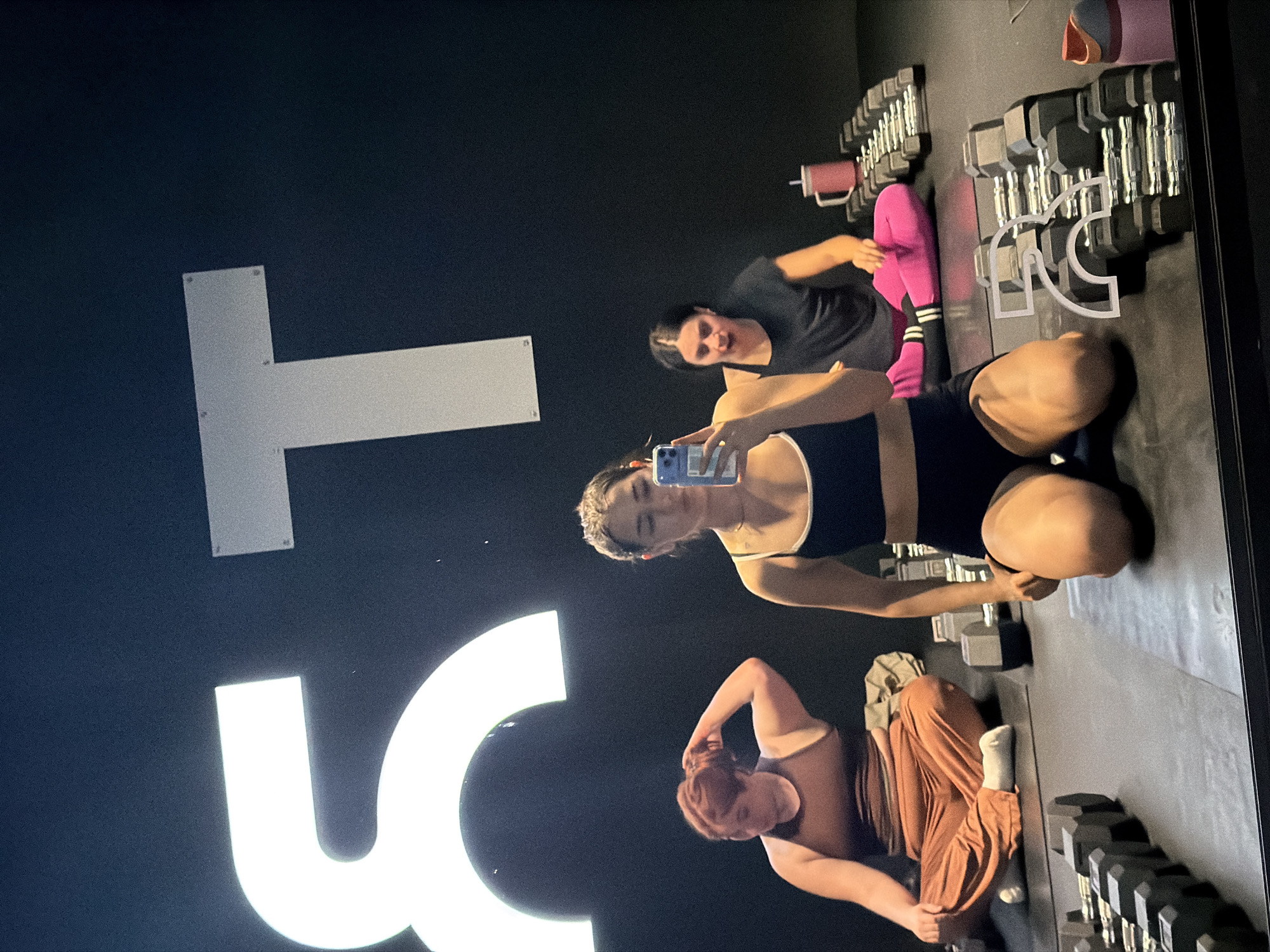 The image shows three women in a gym. The woman in the foreground, seen from the knees up, is wearing a black sports bra and black shorts, and is holding a phone to take a selfie. She is kneeling on a black yoga mat. To her left, a woman with red hair is sitting cross-legged on a mat, wearing a brown tank top and orange pants. She is reaching up to adjust her hair. To the right of the woman in the foreground, another woman with dark hair is sitting with her legs crossed, wearing a black t-shirt and pink leggings with white stripes. She is looking forward and appears to be resting. The gym has a black wall with large white illuminated letters that appear to spell "ST". There are rows of dumbbells on the floor in front of the women, and a pink water bottle is visible near the woman in pink leggings. The image appears to be a reflection in a mirror, as the bottom of the frame shows the edge of a mirrored surface. There is no indication that this image was taken in Puebla, Mexico.